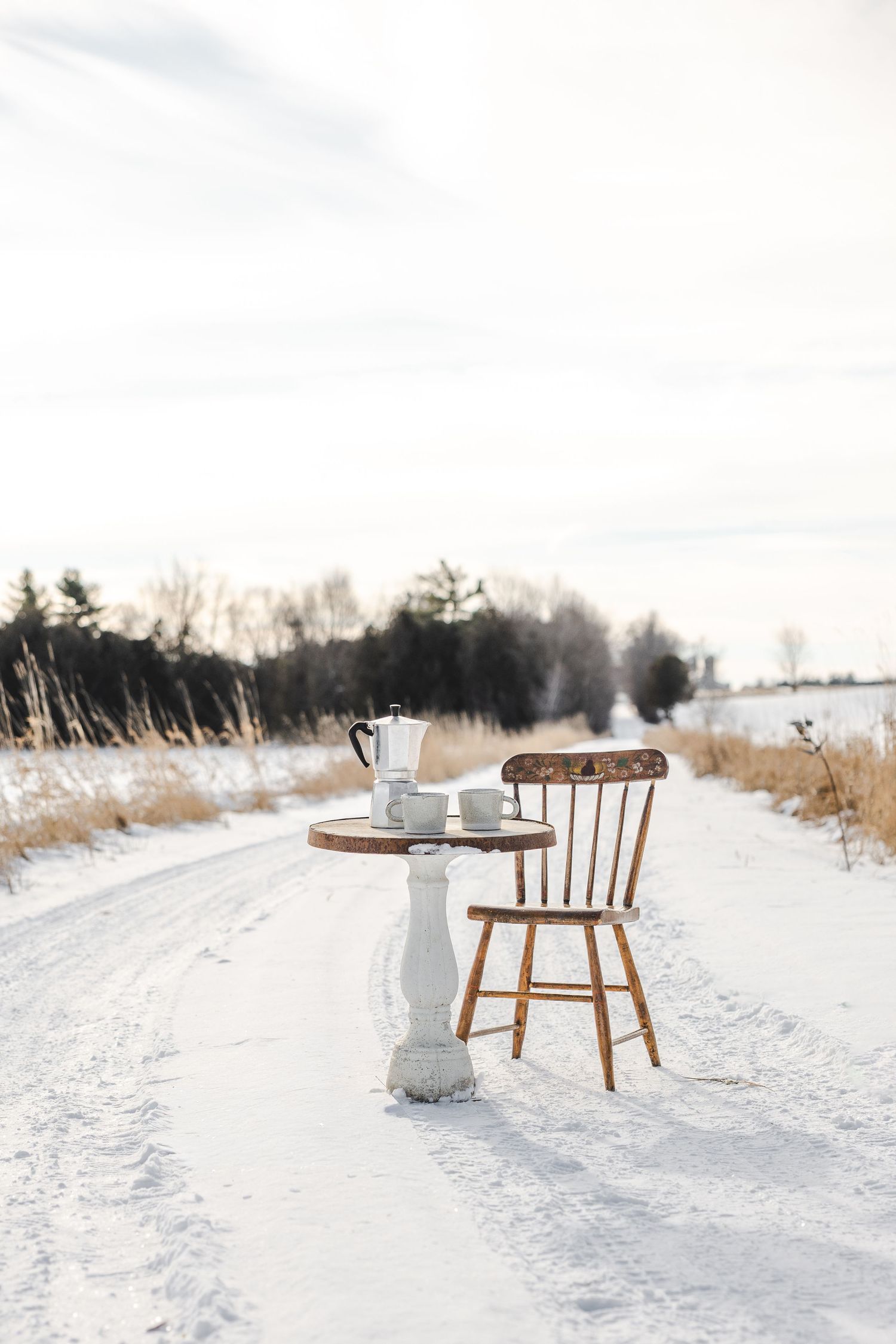 A wooden chair sits alone in a snowy winter landscape with bare trees in the background.