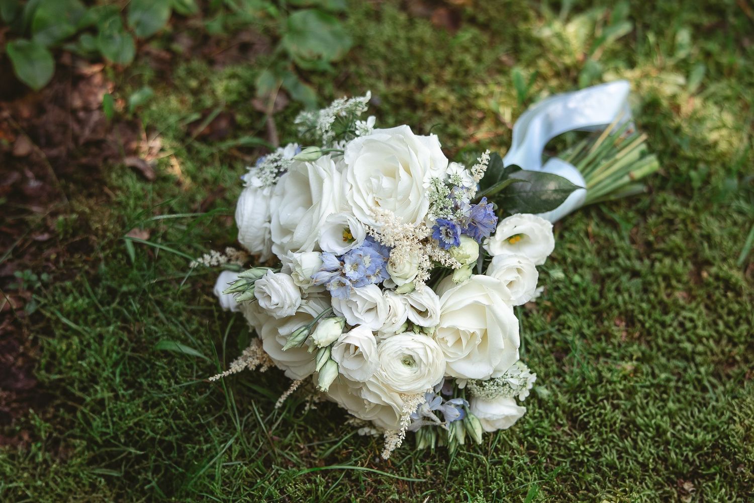 Bouquet de mariage blanc composé de roses et de fleurs délicates, posé sur de la mousse verte en extérieur.