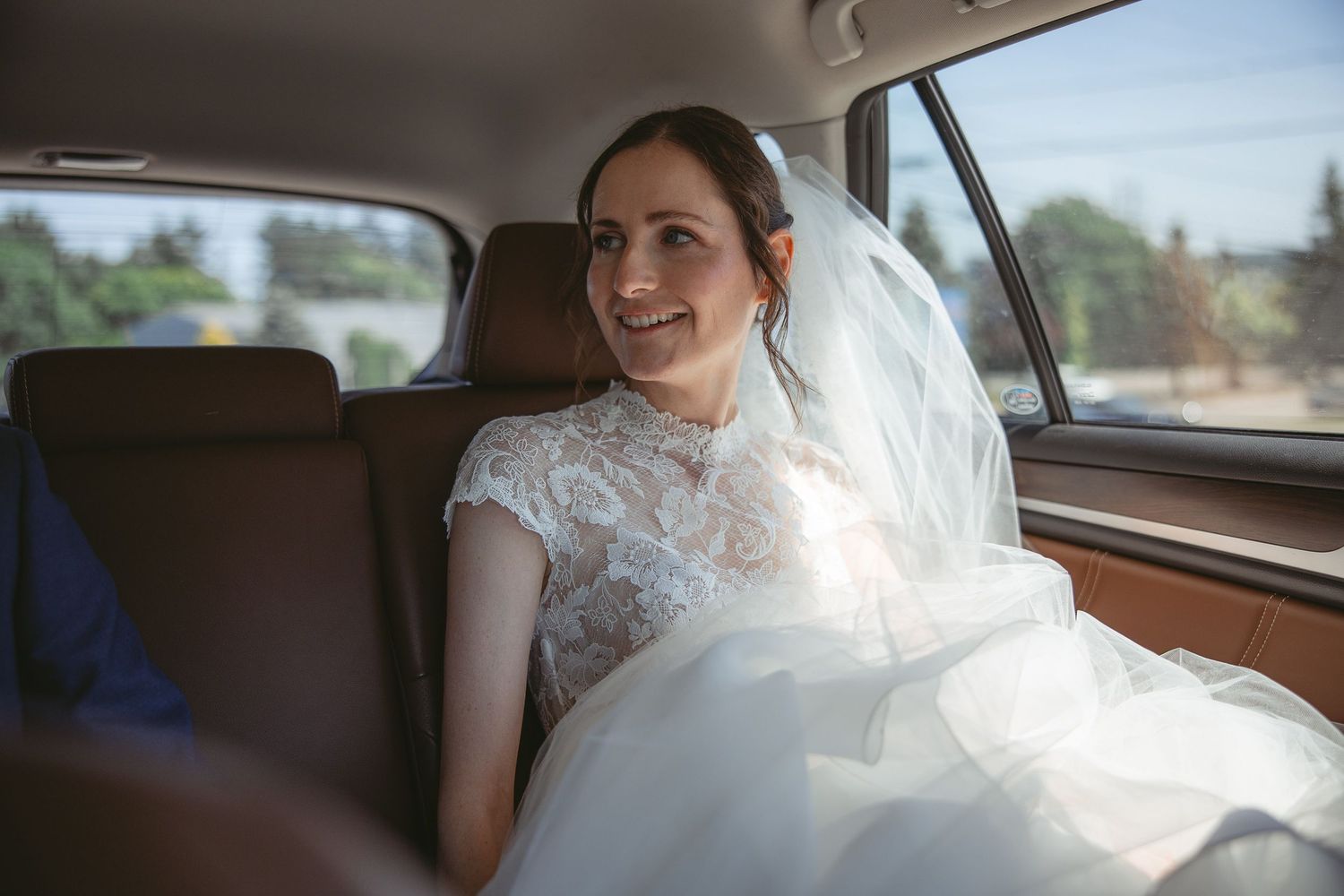 Mariée en robe de mariage blanche en dentelle, assise élégamment à l’arrière d’une voiture vintage lors de la journée de mariage.