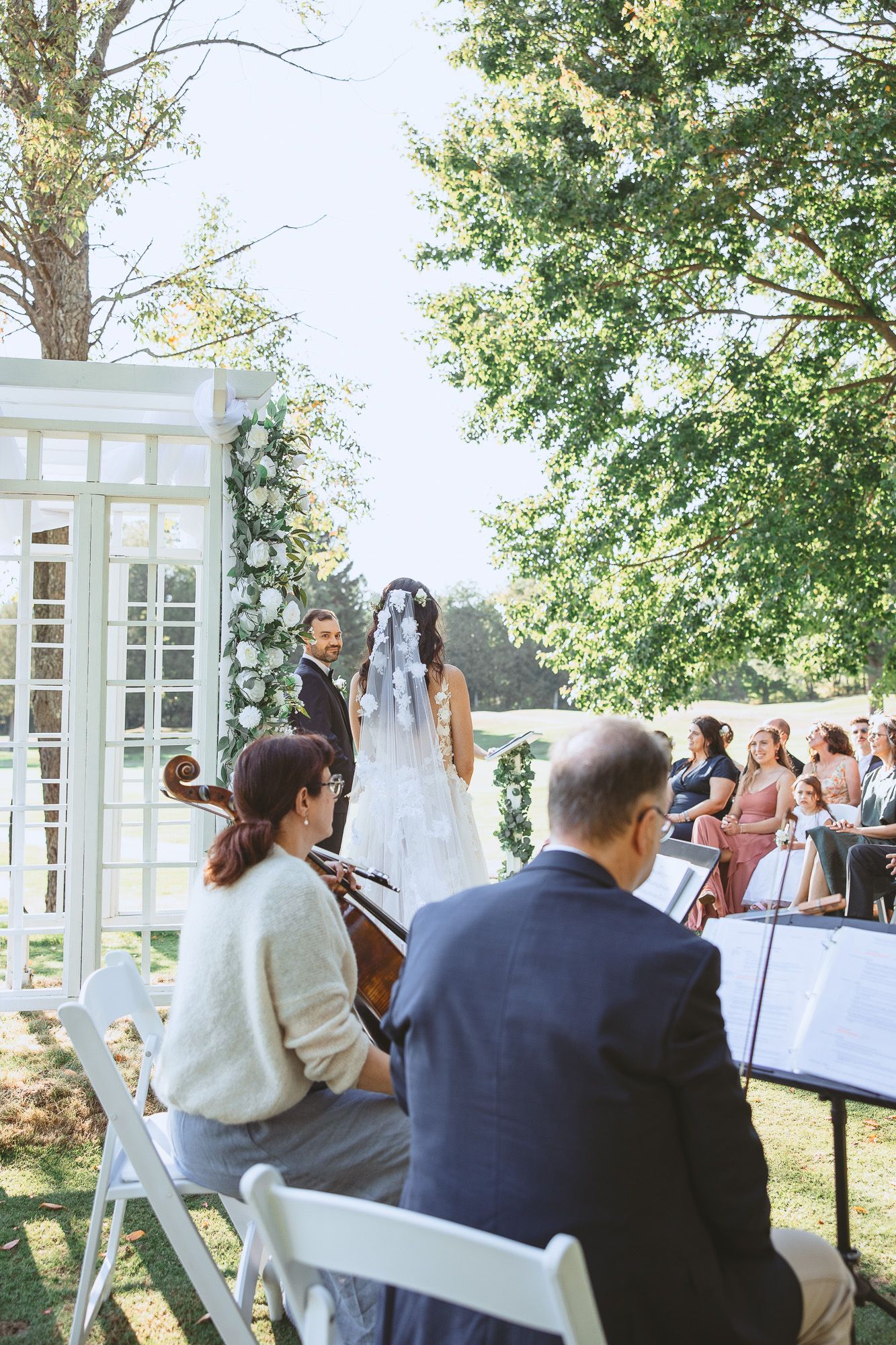 Invités assis lors d’une cérémonie de mariage en extérieur, pupitres de musique devant des panneaux blancs ajourés, journée ensoleillée