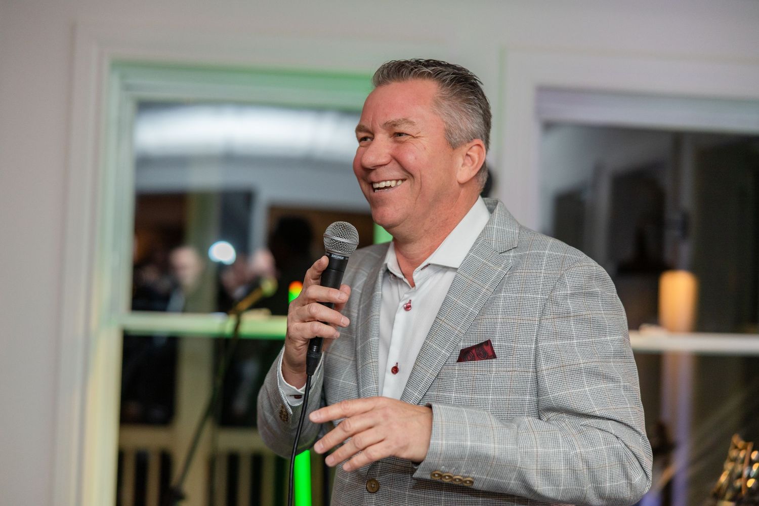 A performer in a light gray suit jacket holds a microphone while speaking or singing at an indoor event.
