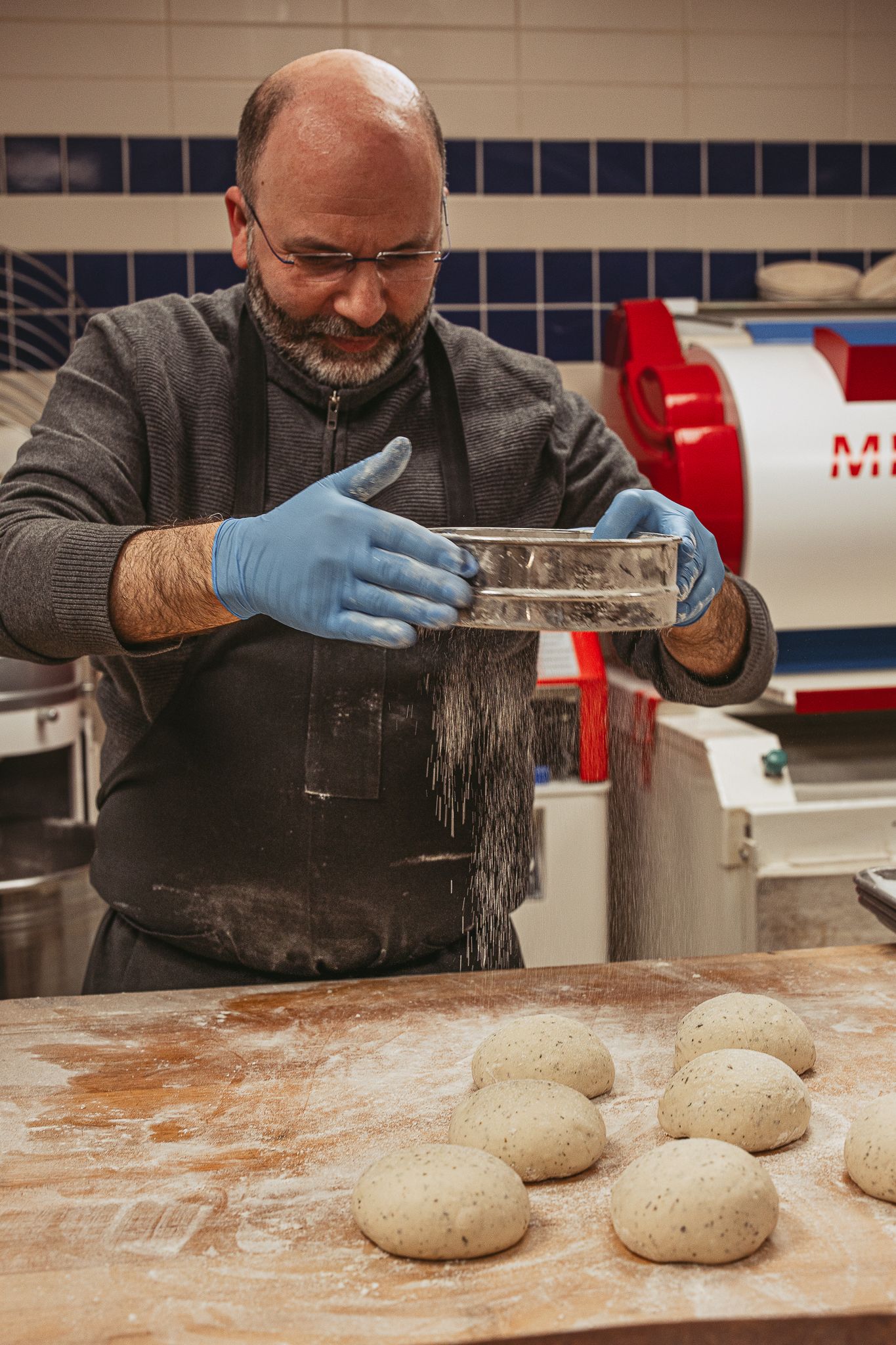 Baker dusting flour over raw bread dough on wooden work surface.