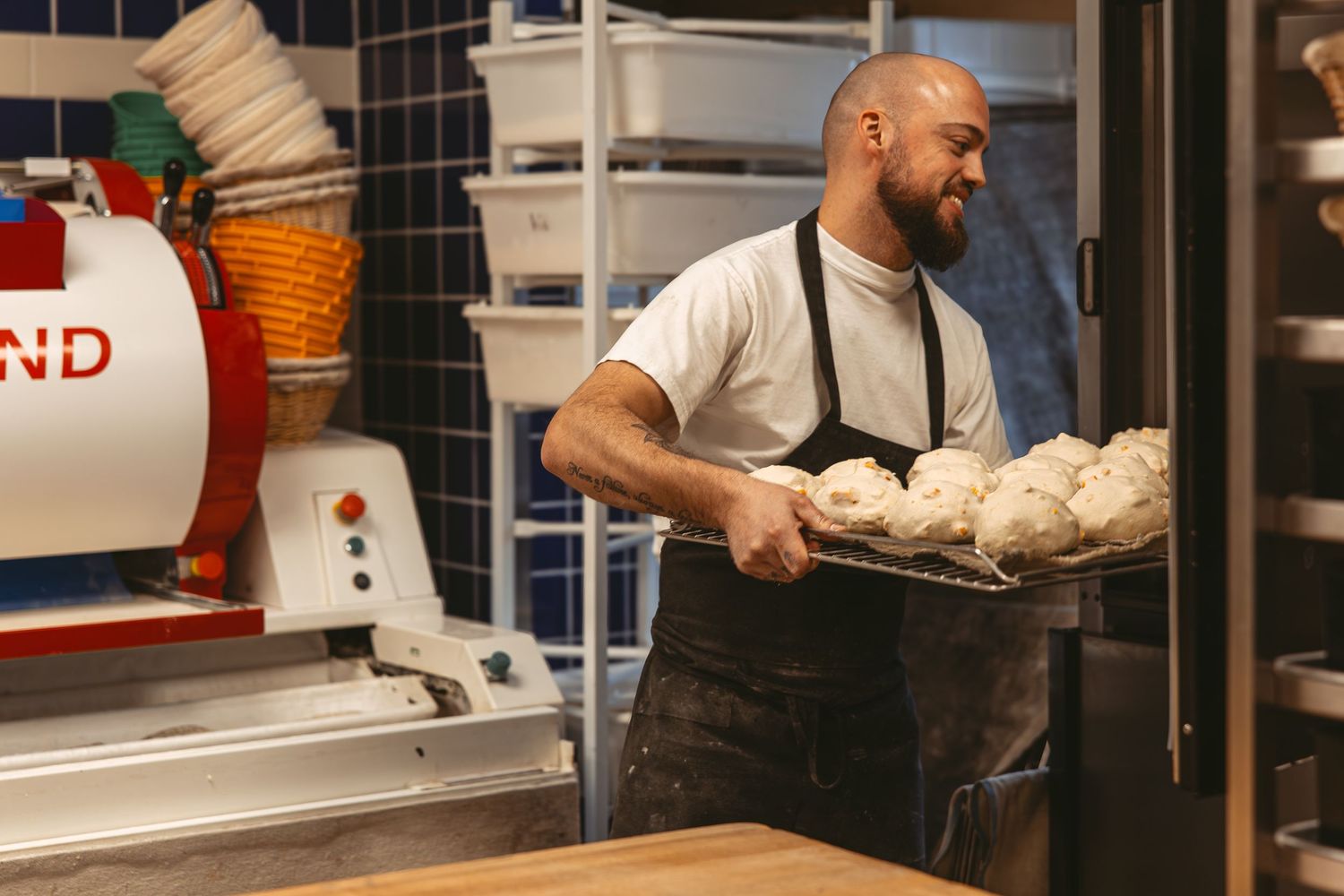 Baker in apron carrying a tray of fresh bread dough in a commercial kitchen.