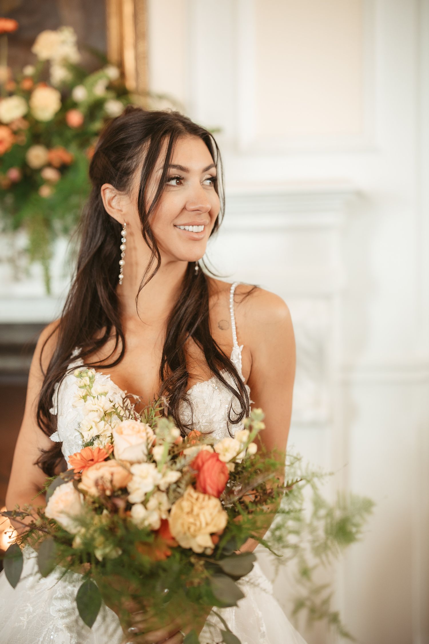 A bride in a white gown smiles while holding a beautiful coral and cream floral bouquet with greenery.