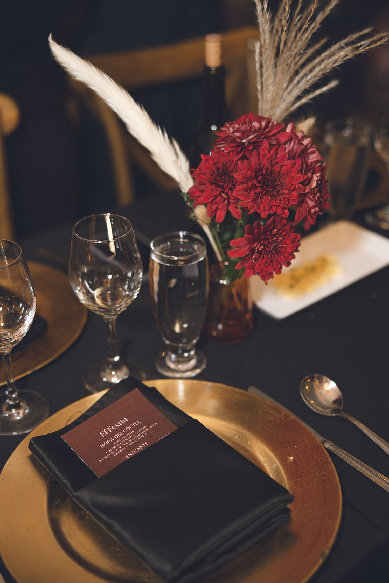 Table de mariage élégamment dressée avec serviettes noires, sous-assiettes dorées, verres à vin et fleurs de carnation rouge foncé.
