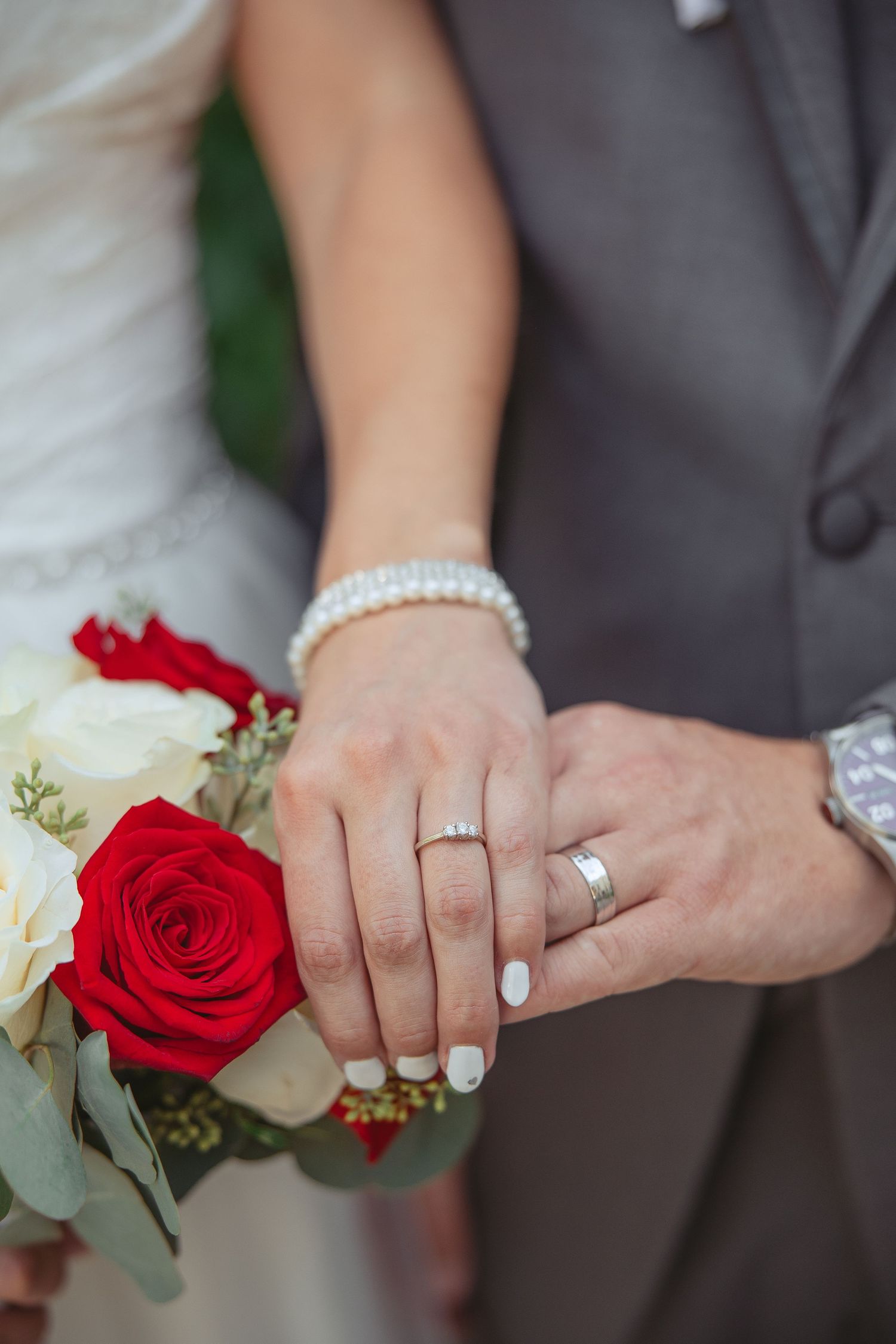 Close-up of newlyweds' hands with wedding rings and red and white rose bouquet details.