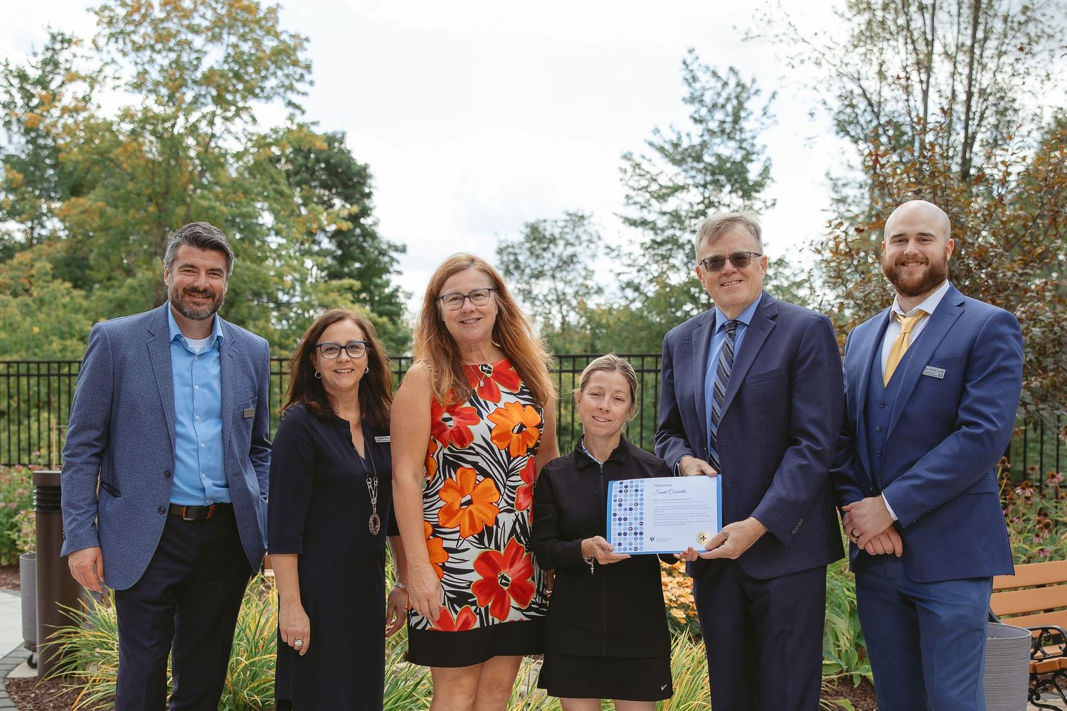 A group of professionally dressed people stand together outdoors near trees and bushes for a photo opportunity.