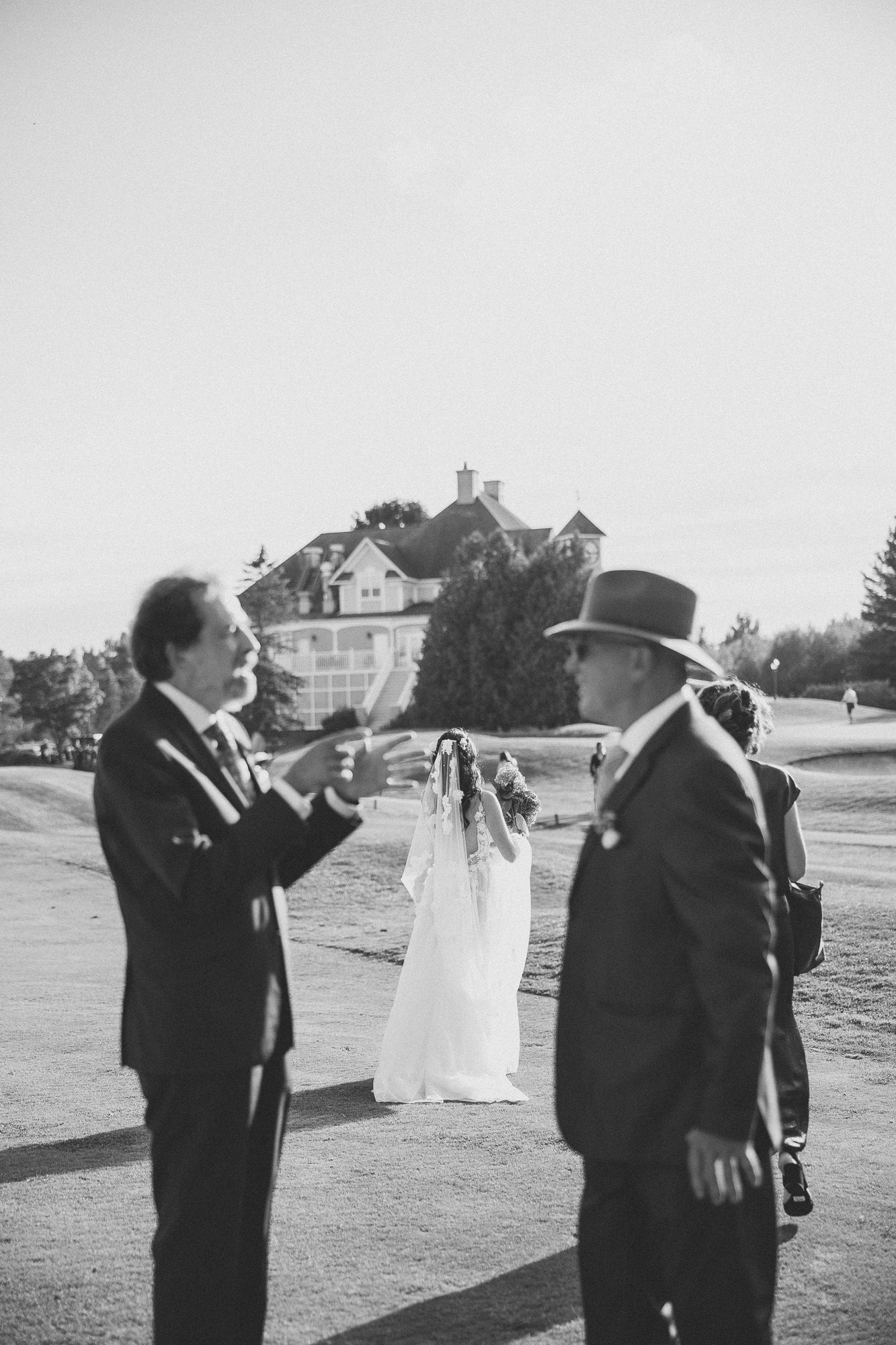 Deux hommes en tenue formelle échangeant en extérieur, avec un grand bâtiment en arrière-plan lors d’un mariage, en Estrie