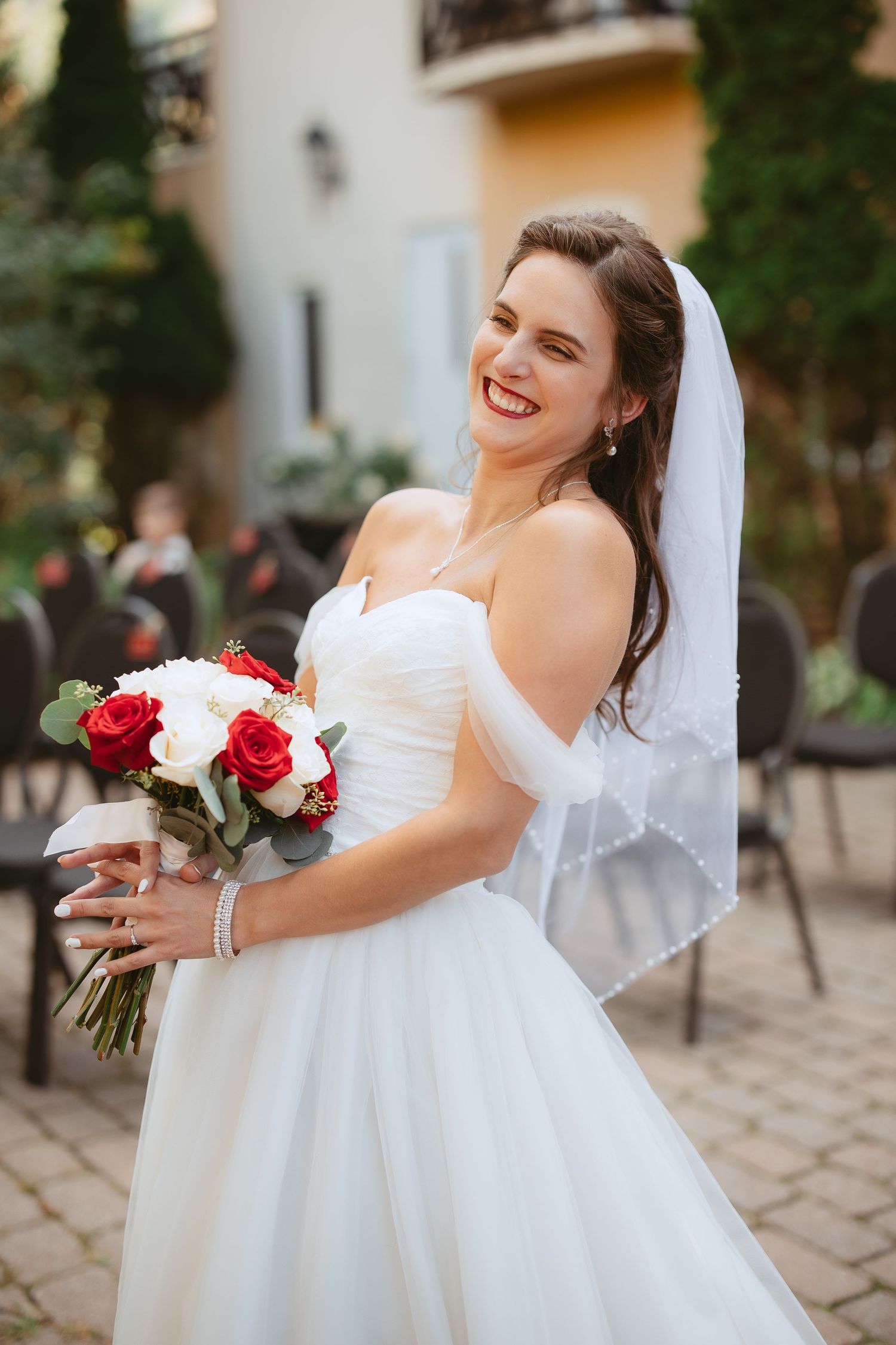 A bride in a white wedding dress holds a red and white rose bouquet while posing outdoors.