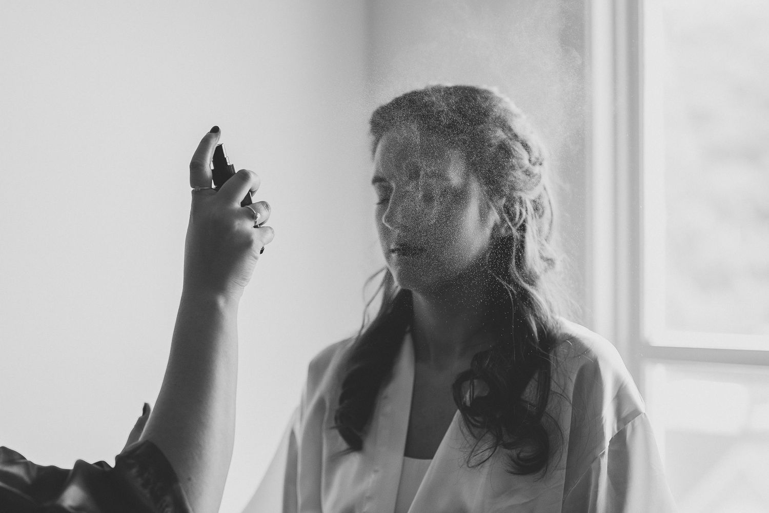 Black and white photograph of someone in a white robe looking at something in their raised hand near a window.