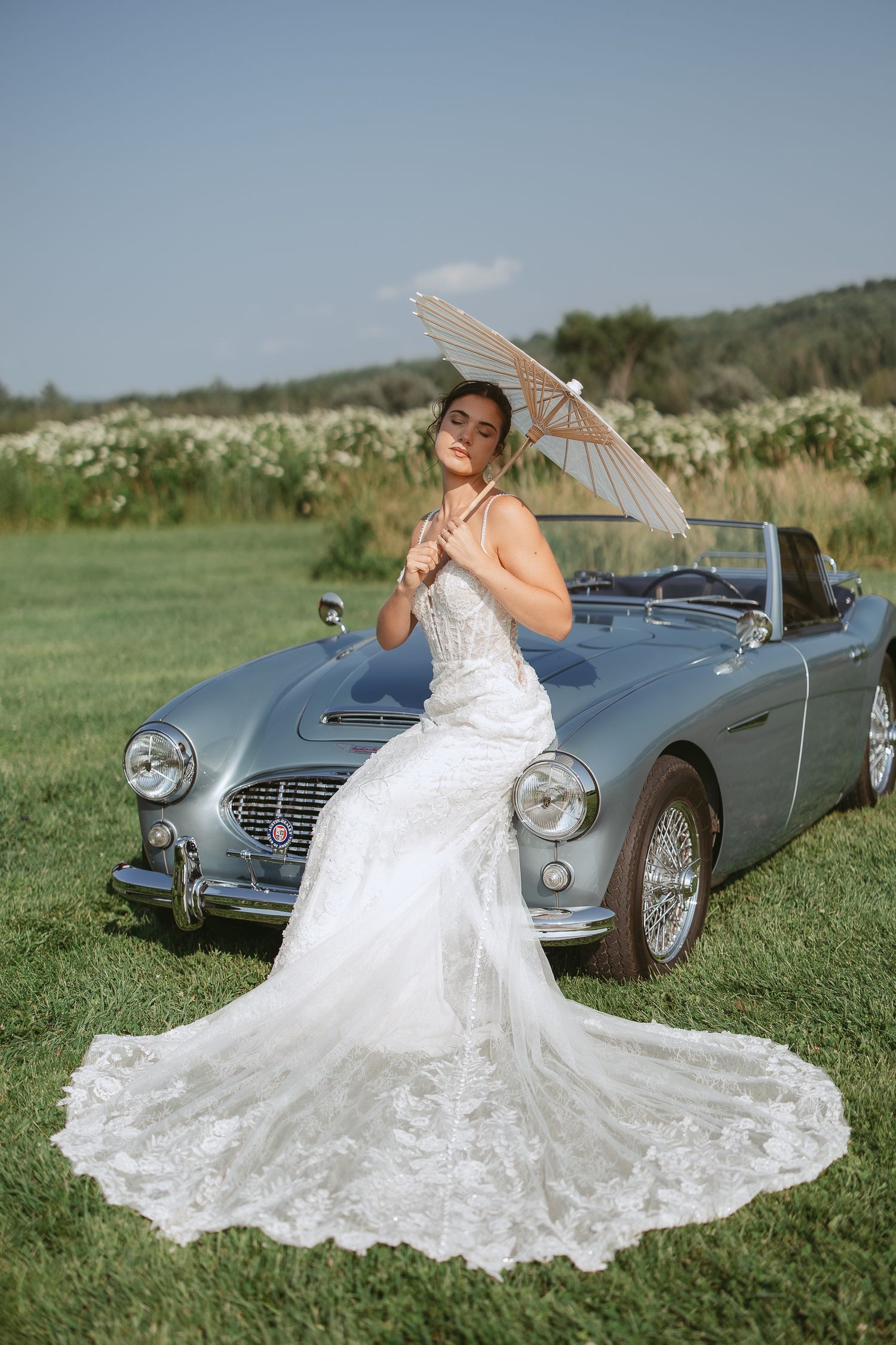 Mariée qui pose avec un parasol appuyer contre une Austin décapotable par une journée d'été