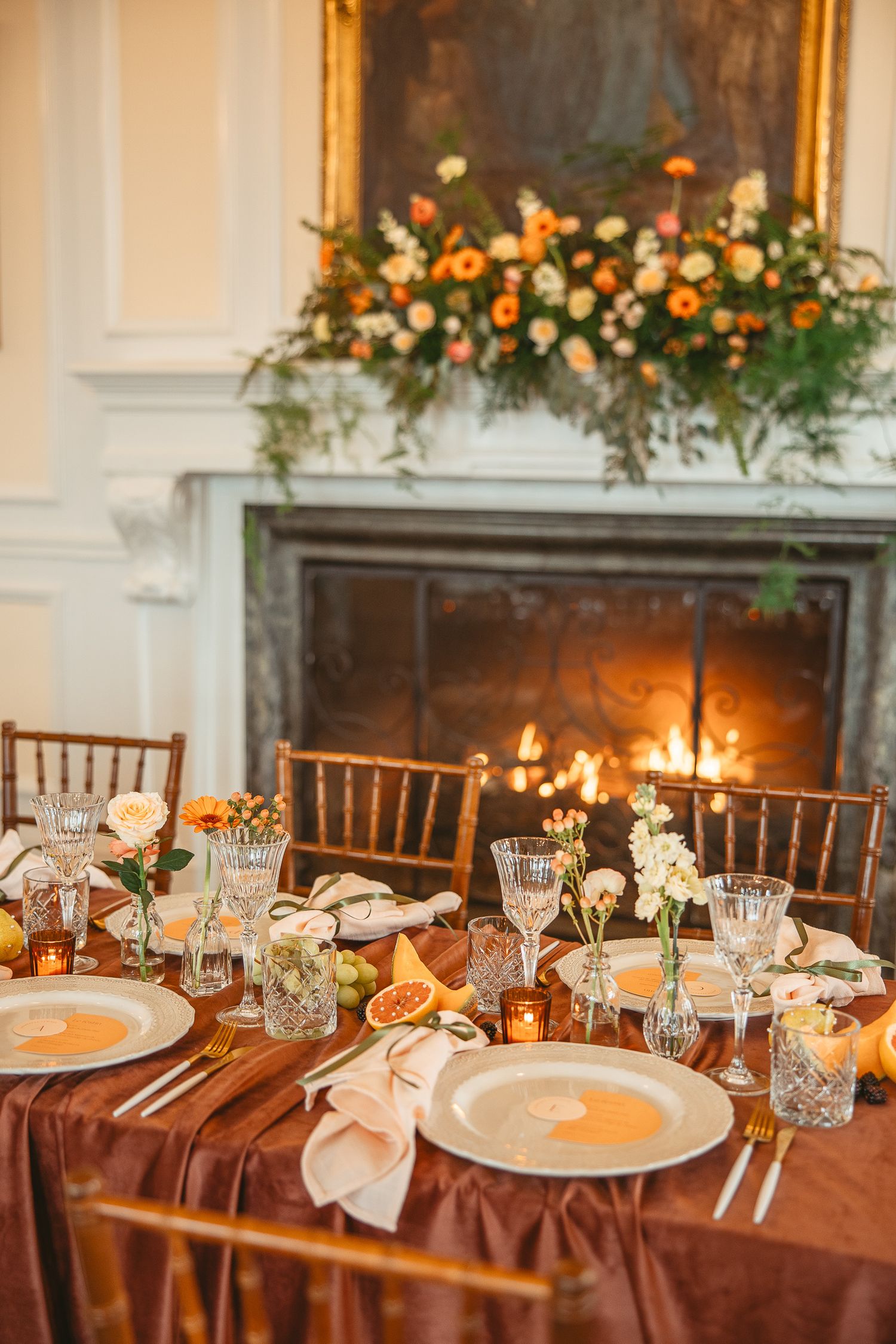 Elegant wedding reception table set with brown linens, crystal glassware, and warm candlelight near a decorated fireplace.