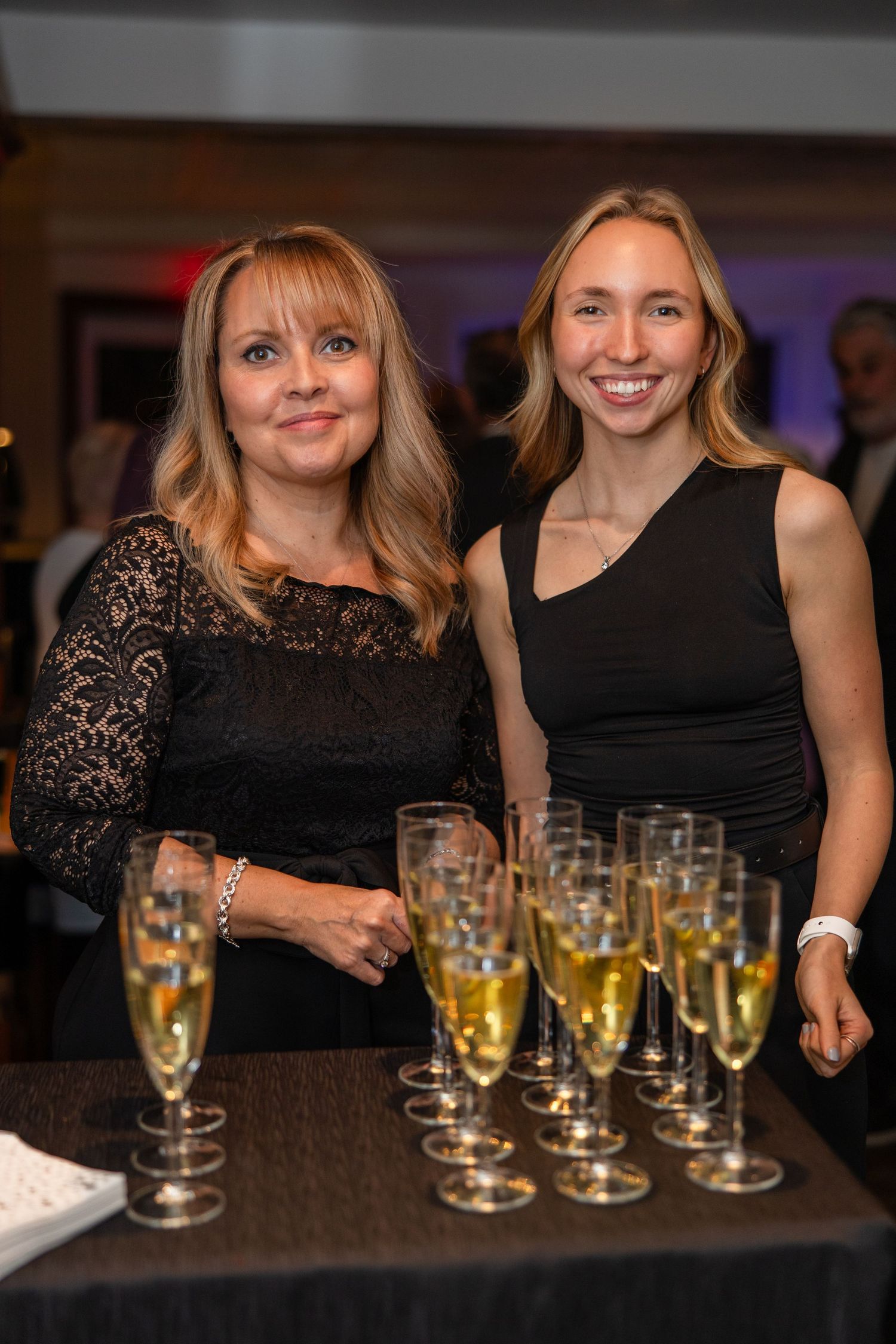 People in black attire gather around a bar counter with rows of champagne glasses during a celebration event.