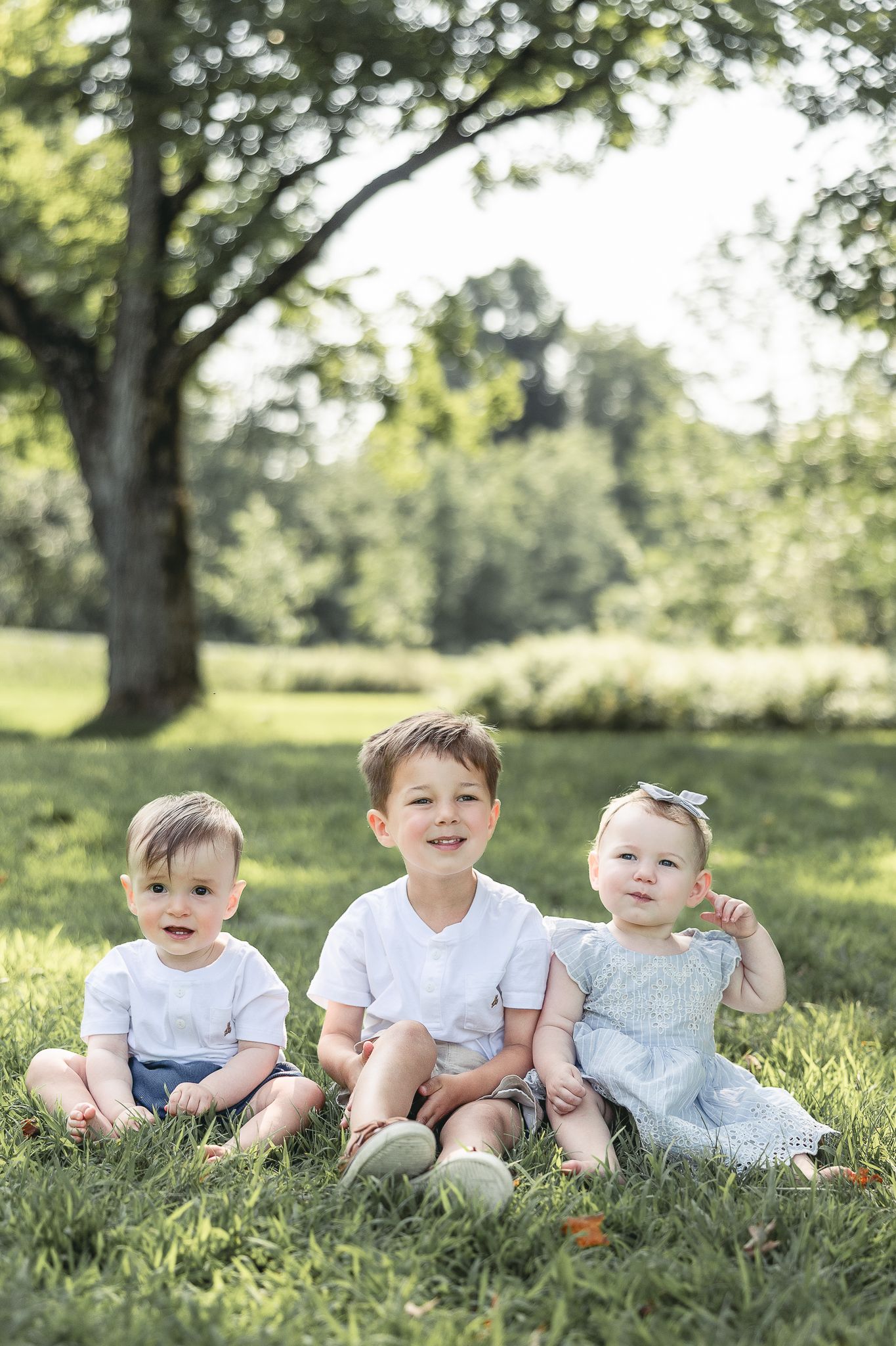 Photographie de trois enfants en tenues claires assis sur herbe verte, captés lors d’une journée estivale ensoleillée, moments authentiques