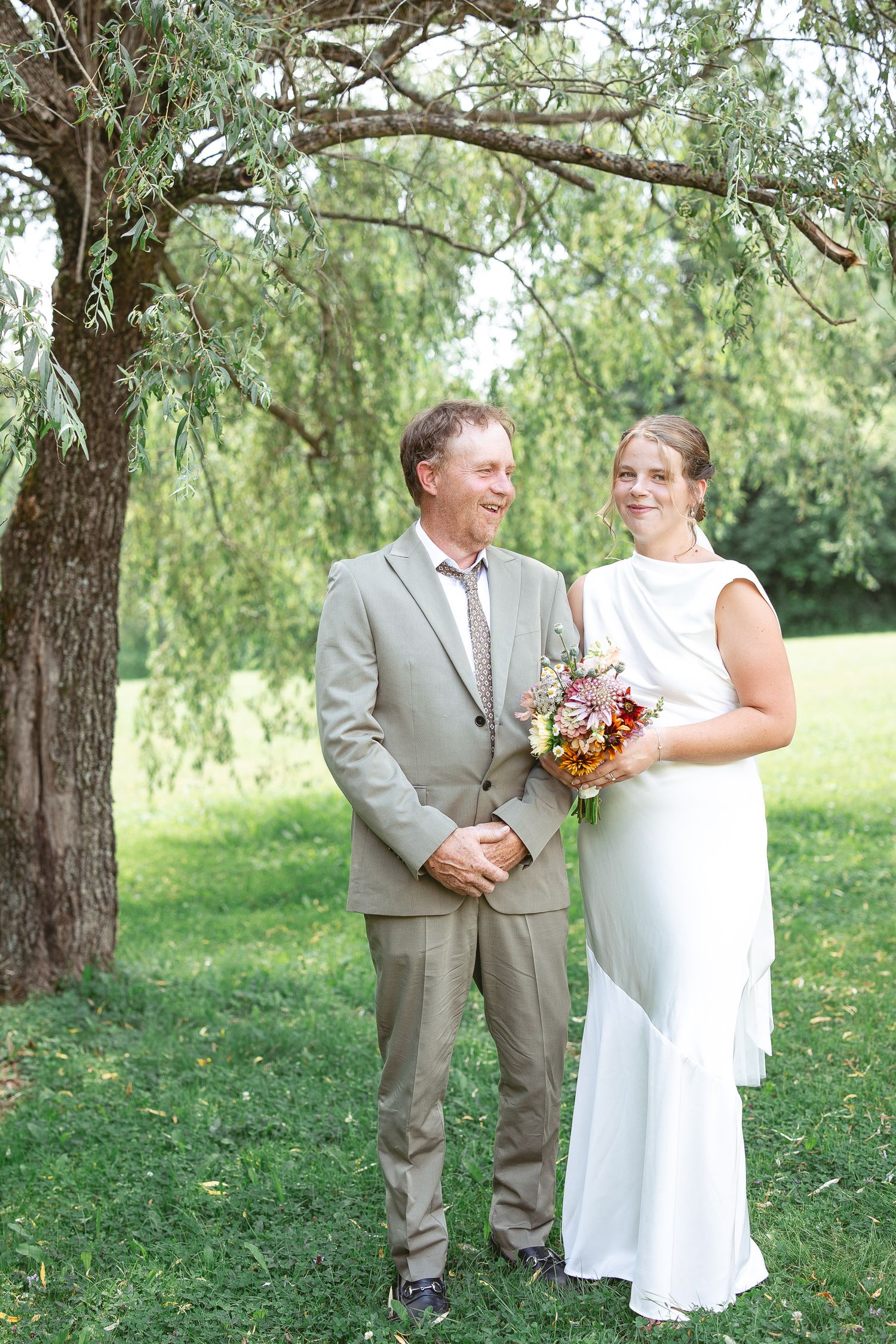 La mariée et son père posant sous un saule par une journée d'été