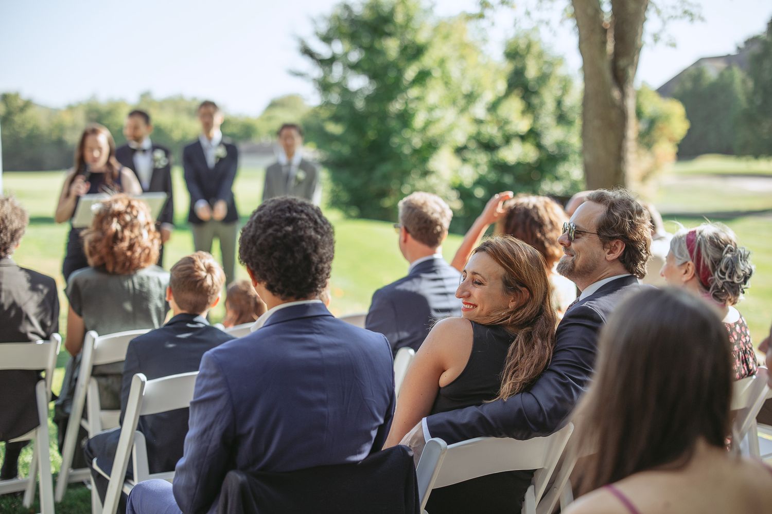 Invités de mariage assis sur des chaises blanches en extérieur, souriant lors d’une cérémonie ensoleillée sur une pelouse