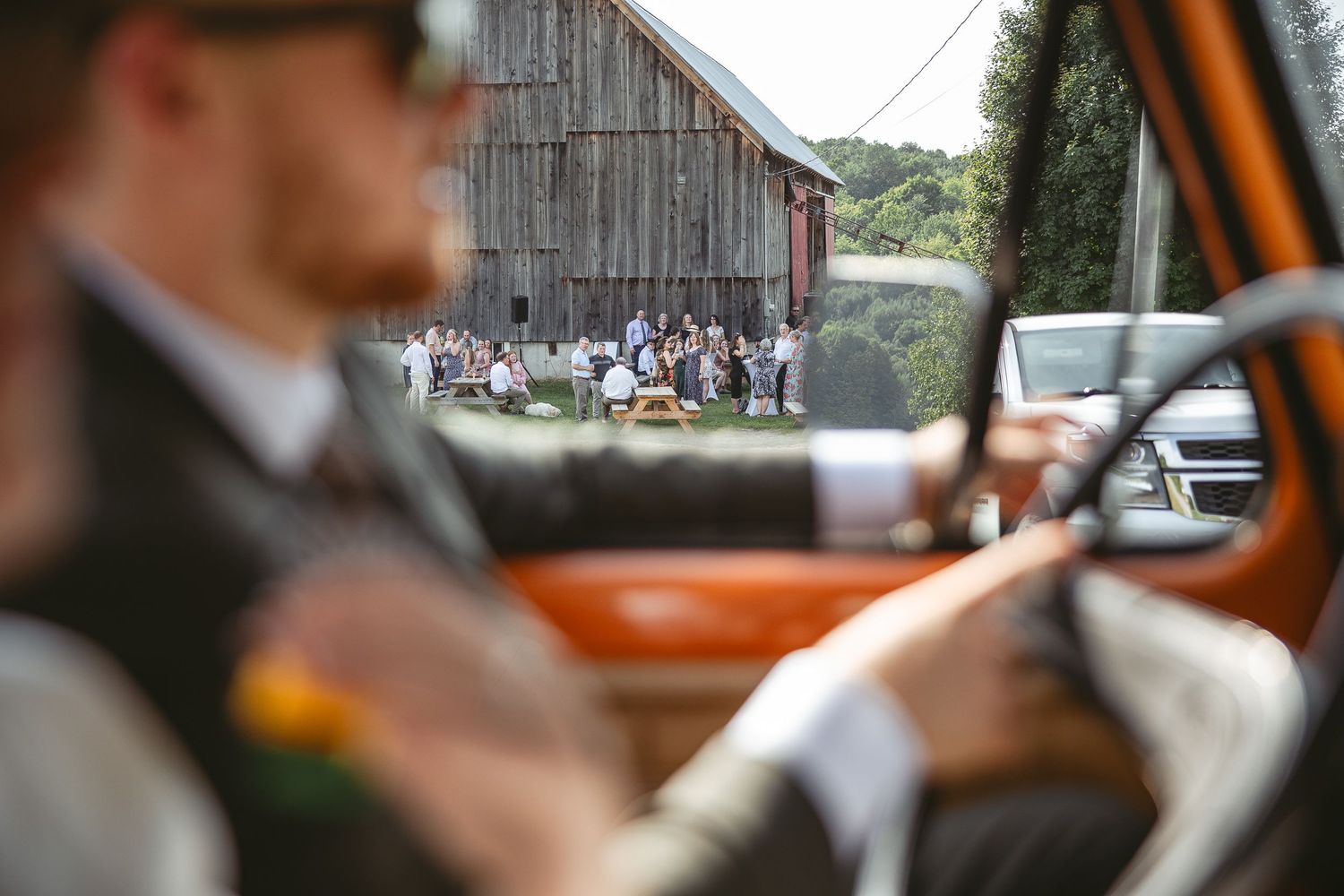 Vue à travers une vitre de voiture d’un rassemblement de mariage rustique, invités réunis devant une grange en bois vieilli
