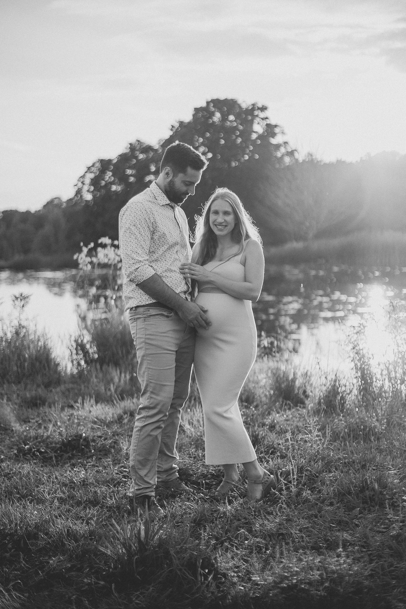 Photographie maternité en noir et blanc : couple au bord d’un lac au coucher du soleil, capturé pour intimité et moments romantiques