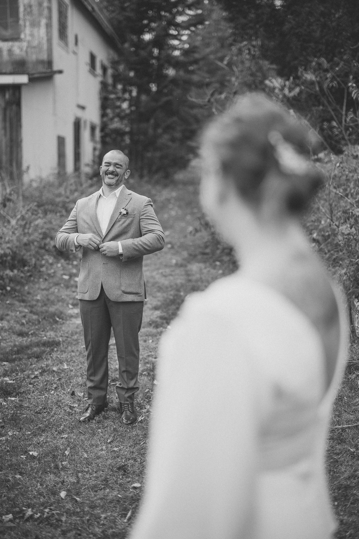 Noir et blanc, les mariés partagent des rires complices en extérieur à l’automne, vêtus d’un complet gris et d’une robe blanche.