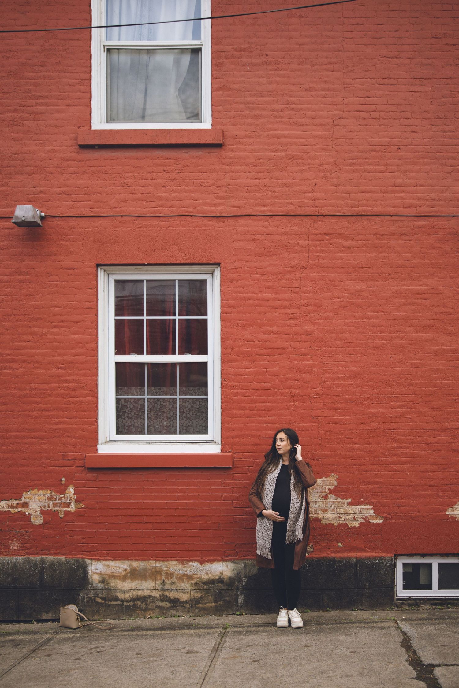Femme enceinte posant devant un mur de briques rouges avec fenêtres blanches, capturée pour mettre en valeur élégance et maternité