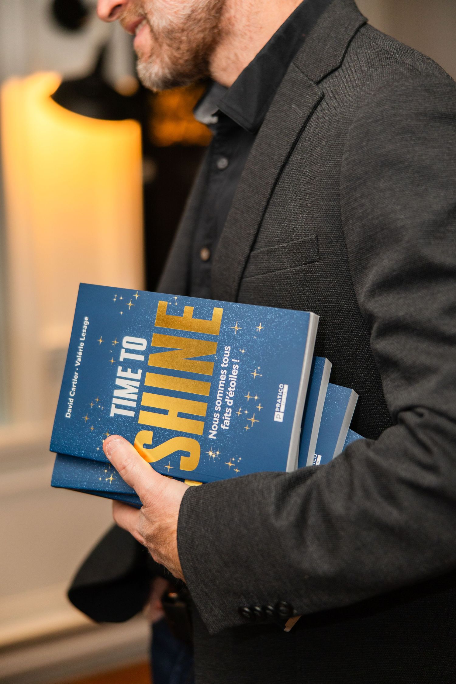 A person in a dark suit jacket holds a blue hardcover book in a warmly lit indoor setting.
