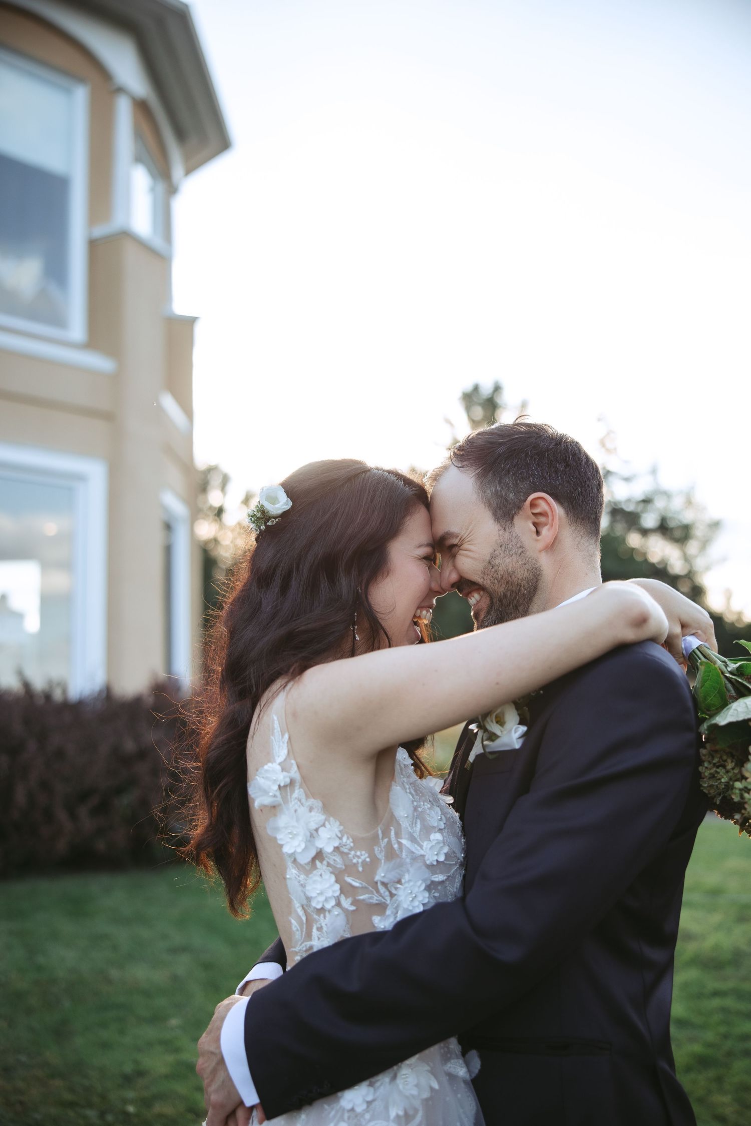 A romantic wedding couple embraces outdoors near a building with large windows at sunset.