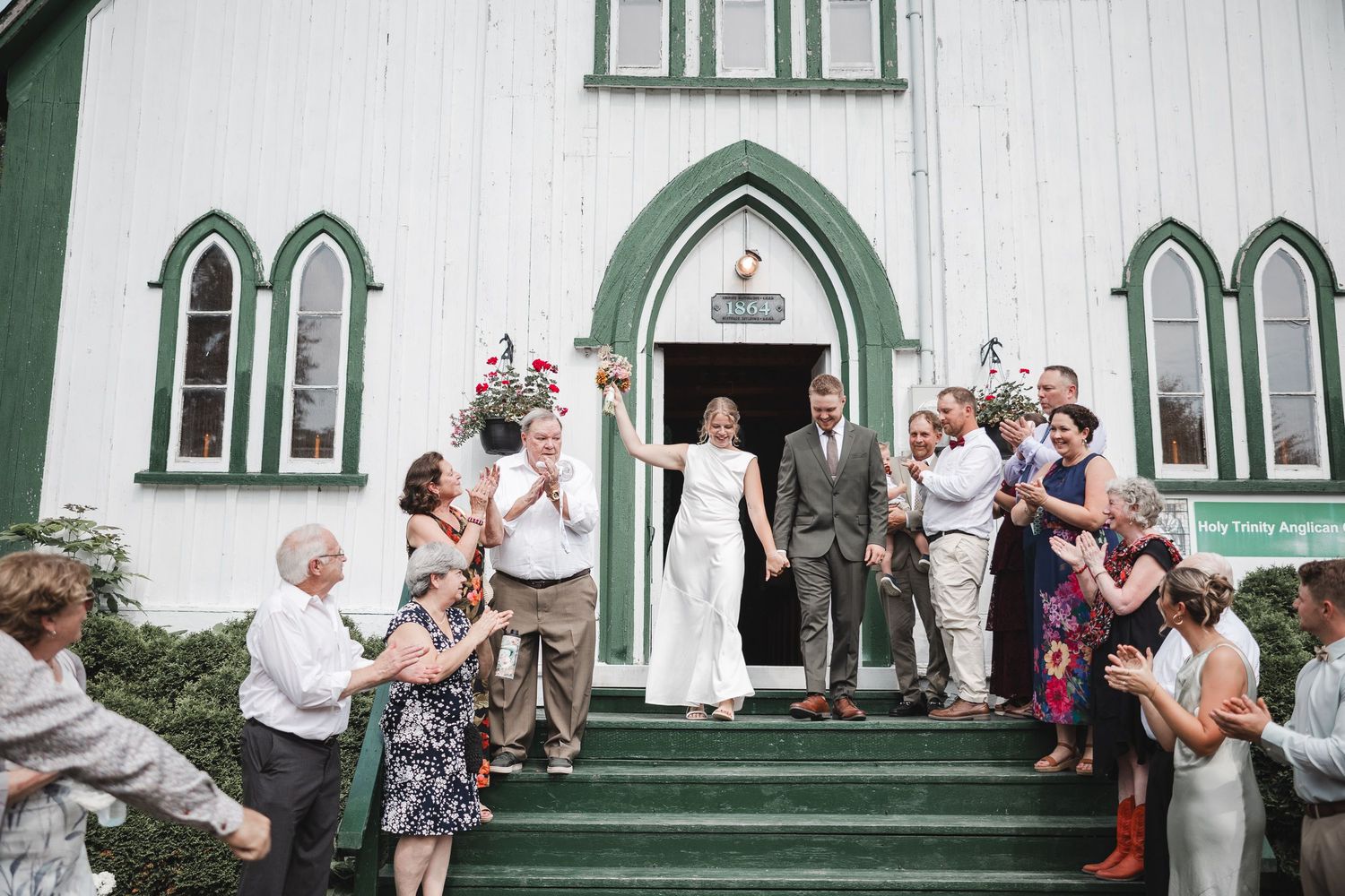 Sortie des maries et leurs invités de mariage devant une chapelle blanche aux fenêtres gothiques vertes et porte voûtée