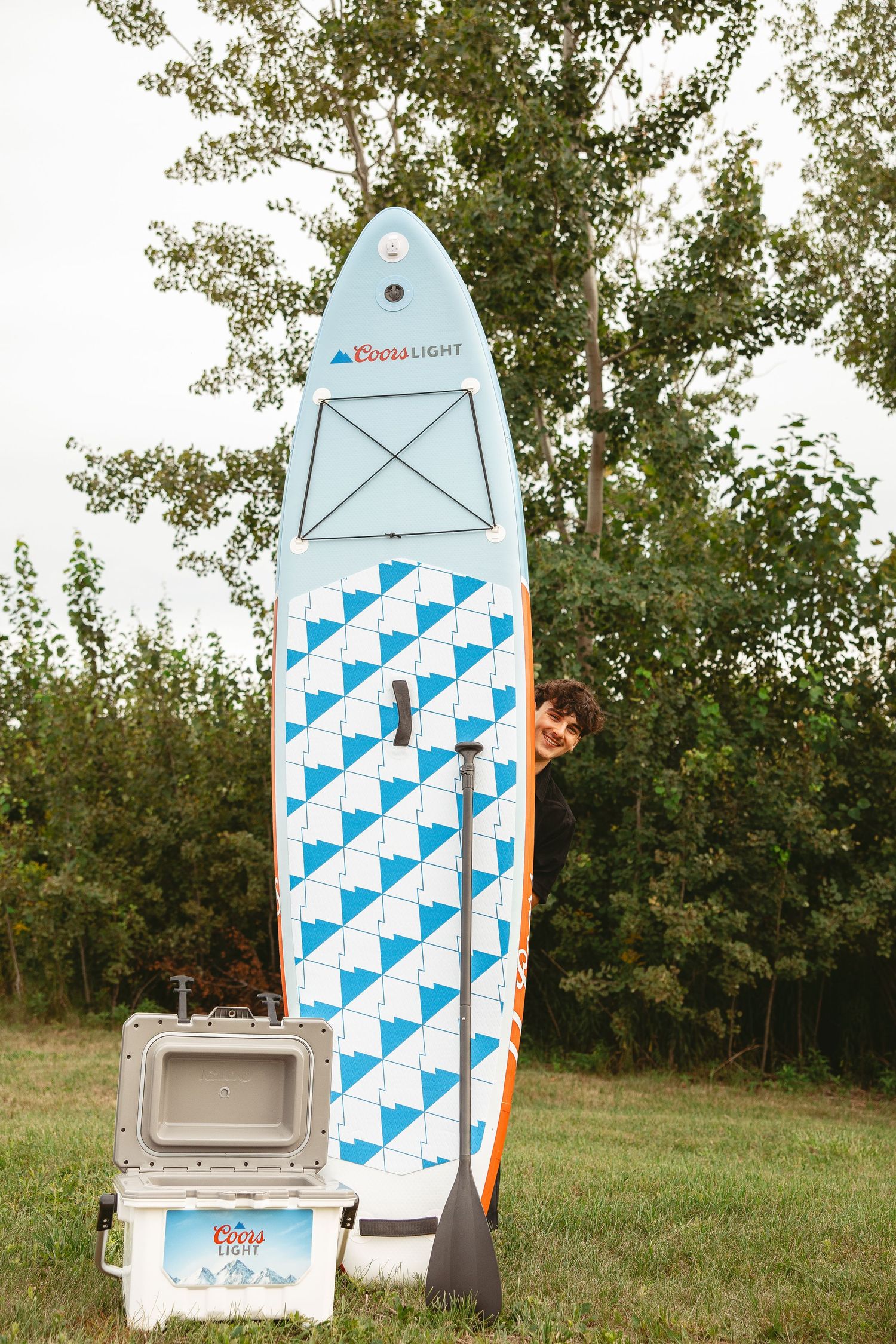 A row of white and blue patterned paddleboards displayed outdoors on grass with coolers nearby.