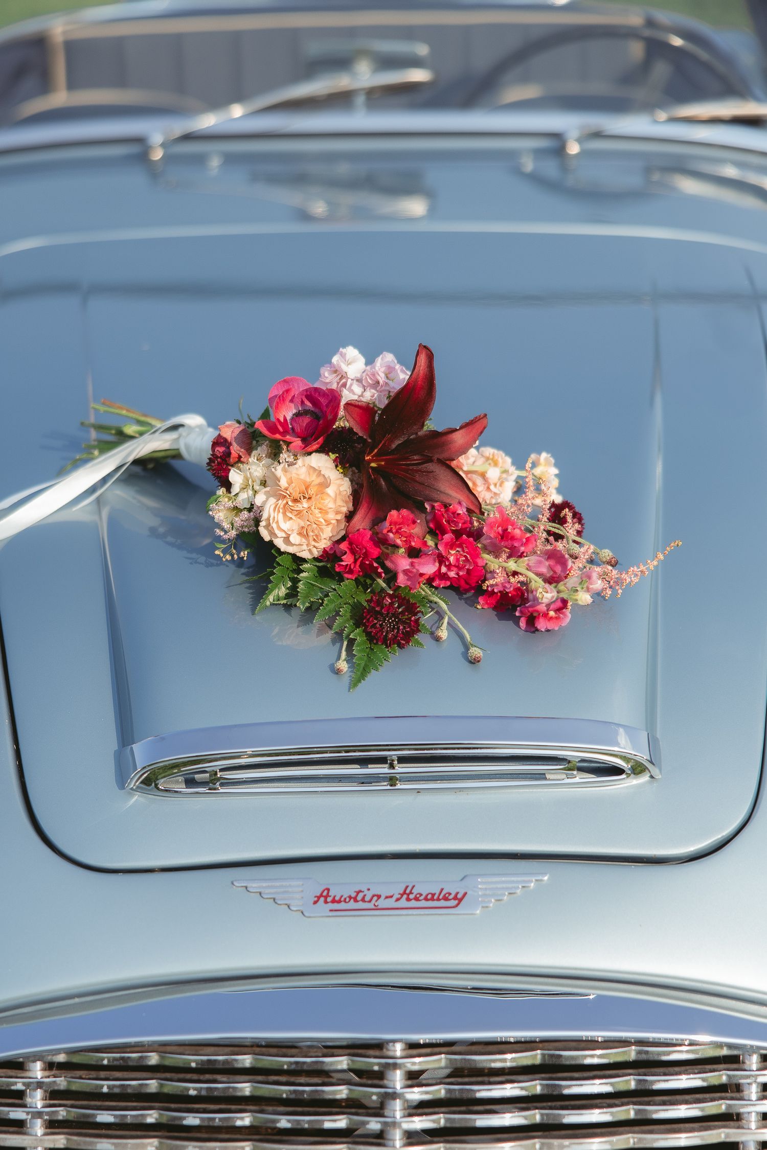 Capot d’une Austin Healey vintage décoré de fleurs roses et pêche, mettant en valeur la calandre chromée lors d’un mariage.