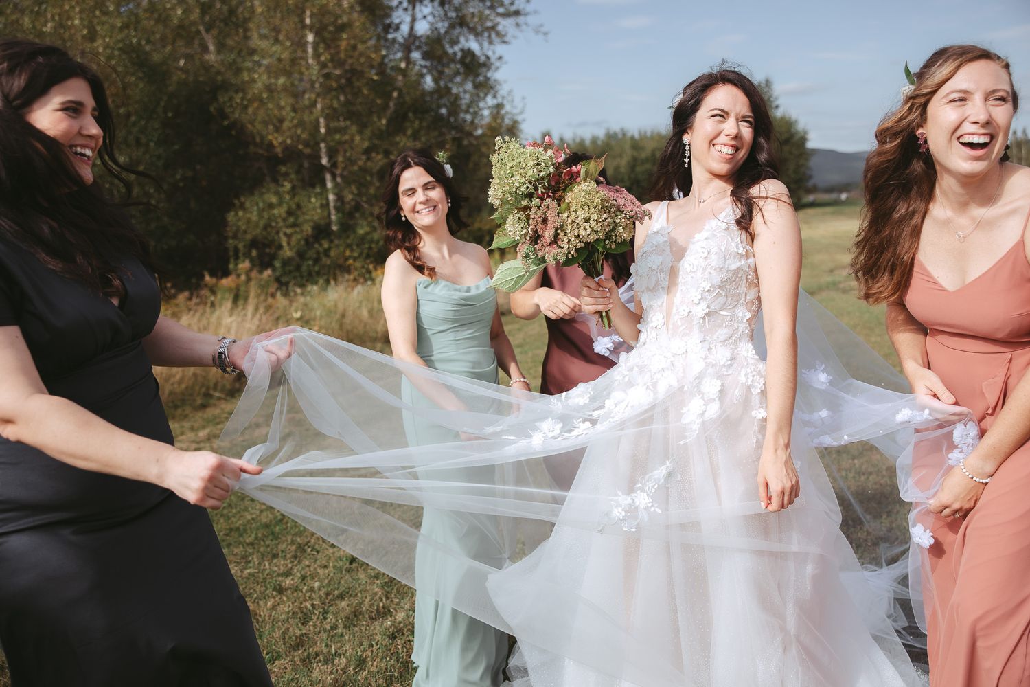 Demoiselles d’honneur en robes pêche ajustant un voile de mariée fluide en extérieur par une journée ensoleillée, en Estrie
