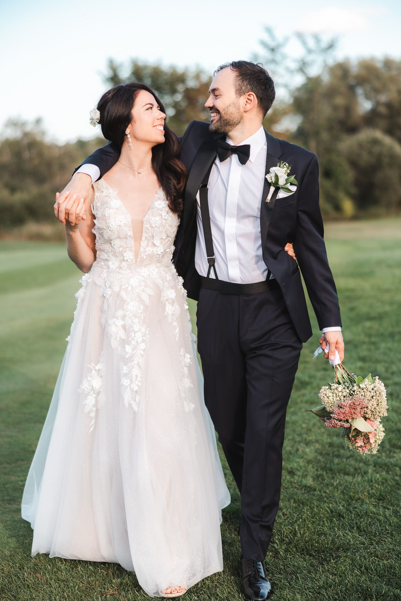 Couple en tenue de mariage partageant un moment joyeux en marchant sur l’herbe, mariée en dentelle et marié en tuxedo, en Estrie