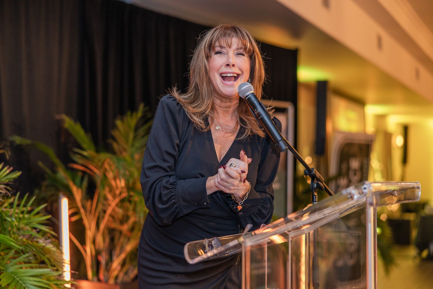 A speaker gestures while delivering a presentation at a podium during an indoor event with warm lighting.
