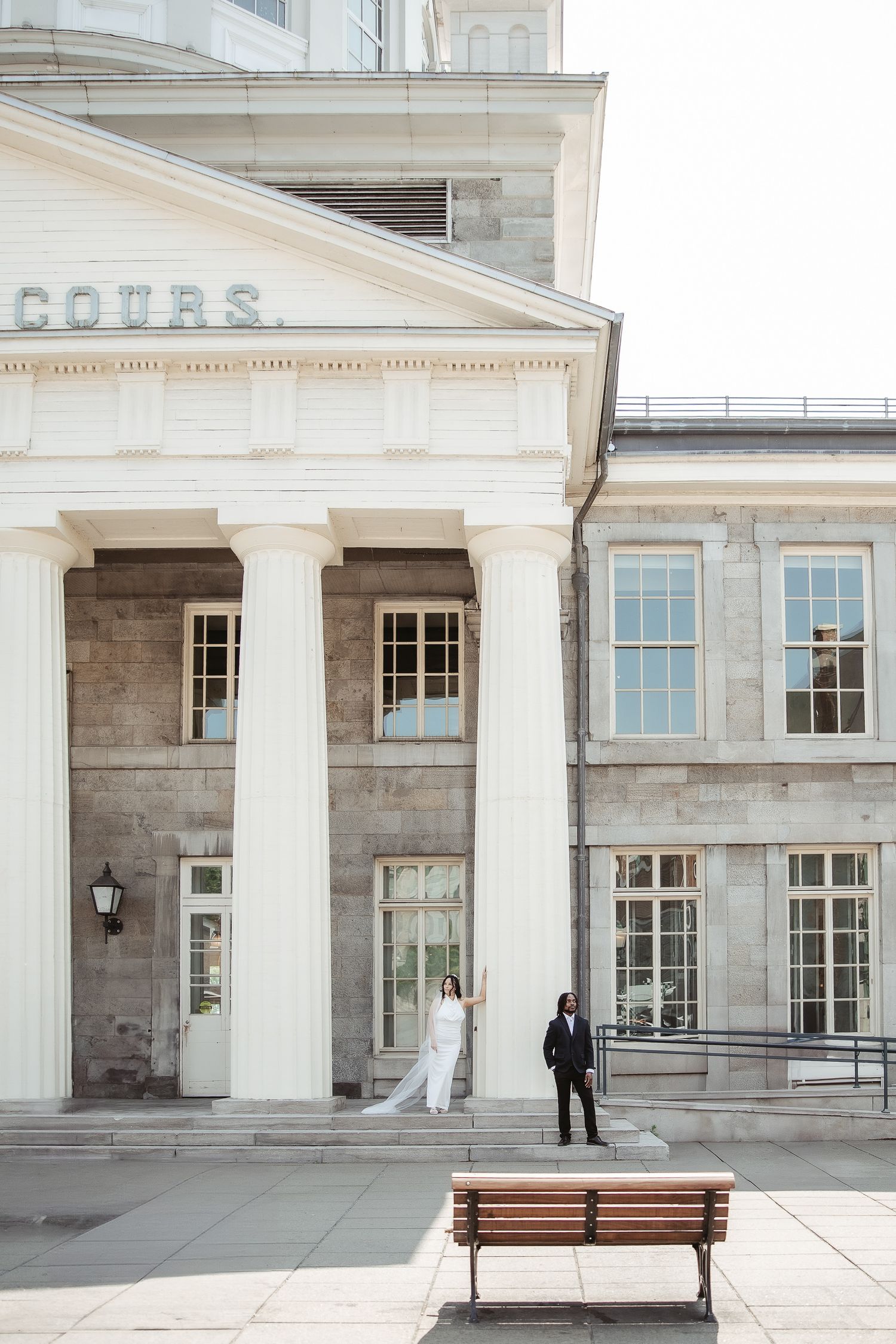 Mariés dans le Vieux-Montréal mettant en valeur la façade d’un bâtiment classique avec colonnes, fenêtres et bancs en bois à l’avant
