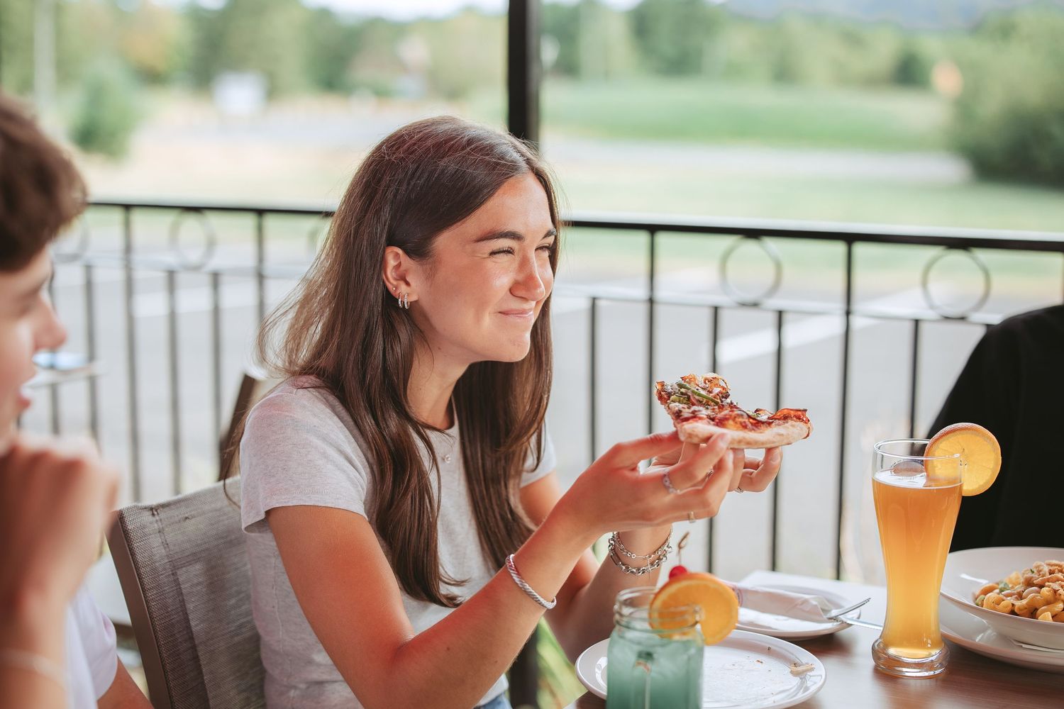 A woman enjoying pizza and beer on an outdoor restaurant patio during a sunny afternoon.