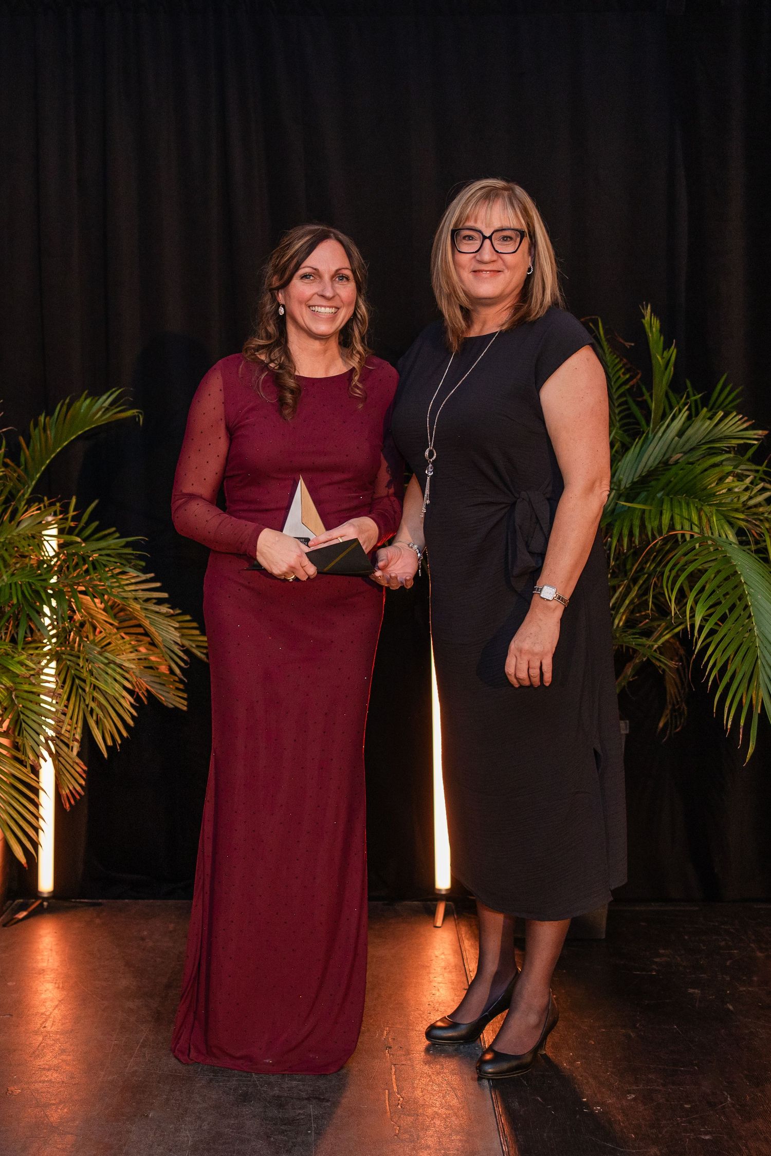 Two women pose together at formal event with palm plants and dramatic lighting in background.