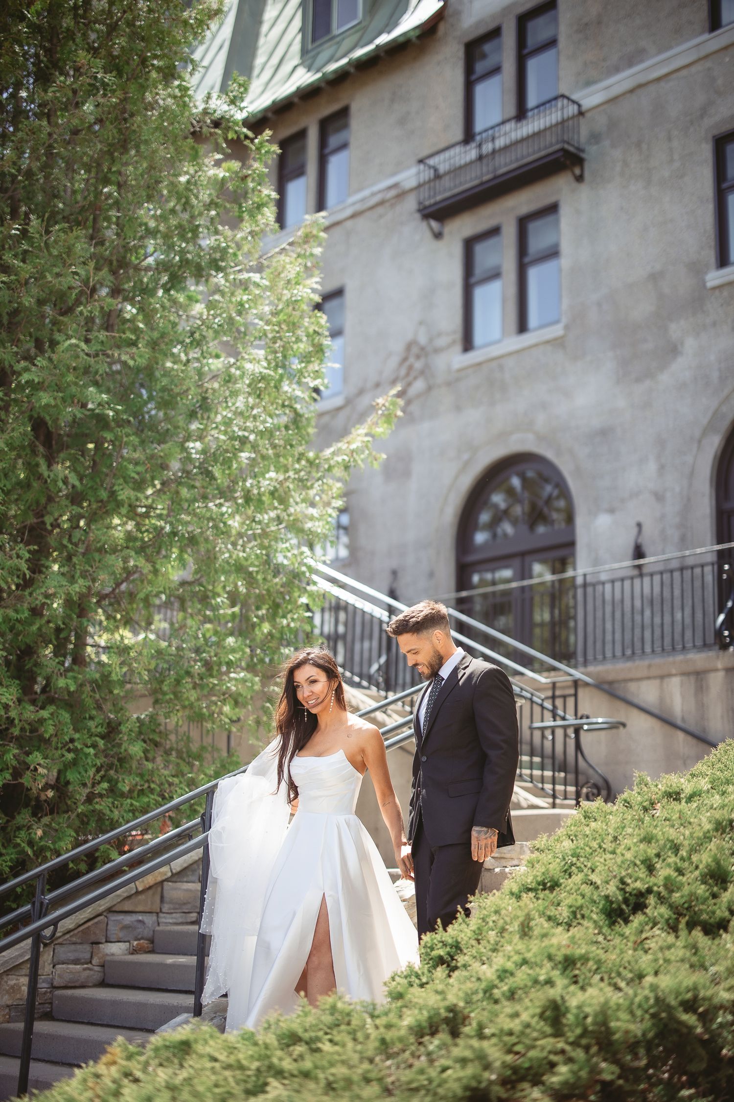 Un couple en tenue de mariage monte des marches de pierre, entouré d’une végétation luxuriante, à l’extérieur d’un bâtiment historique.
