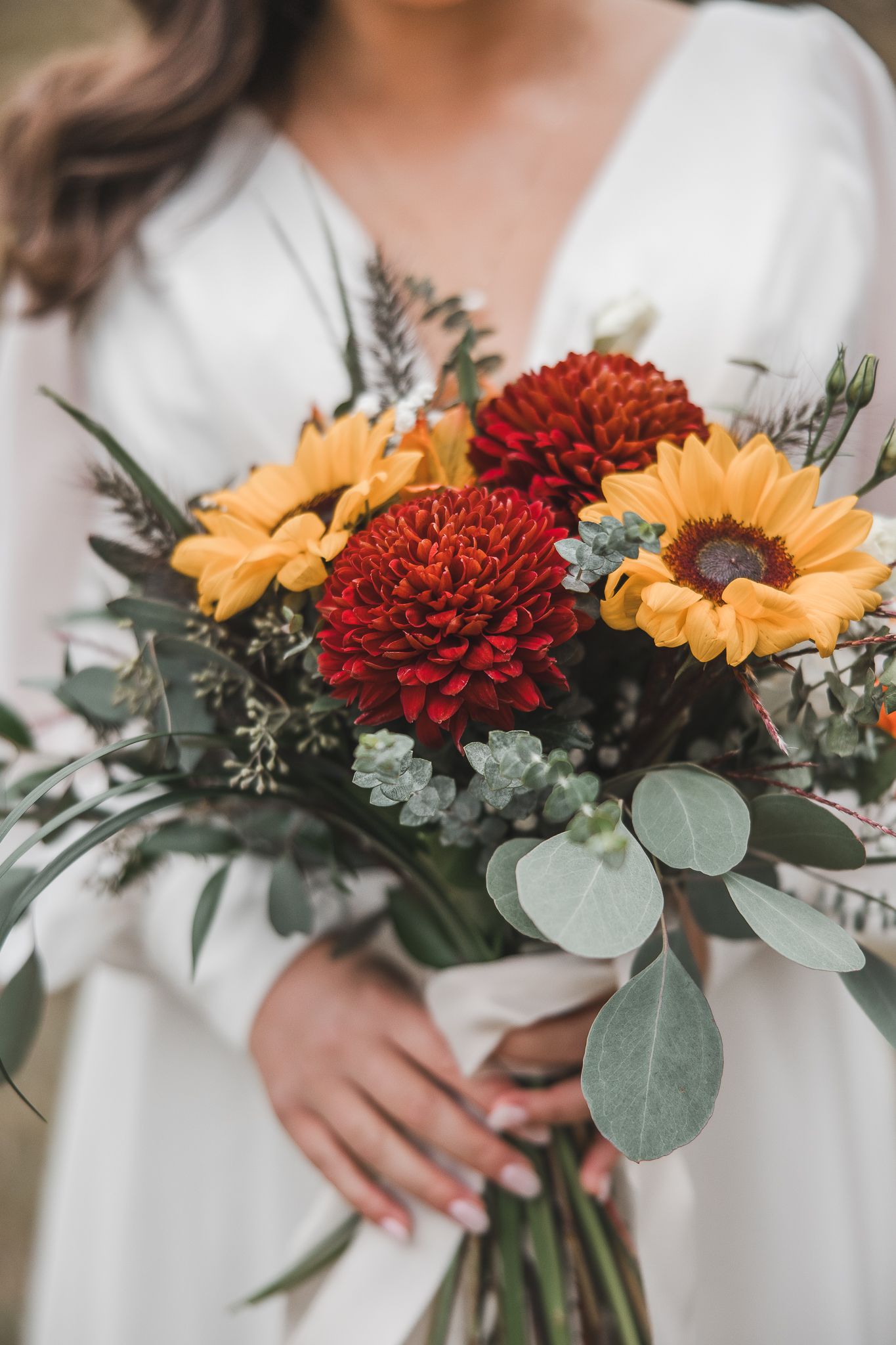 Un bouquet de mariée composé de dahlias bourgogne, de tournesols jaunes et de feuilles d’eucalyptus, tenu contre une robe blanche.