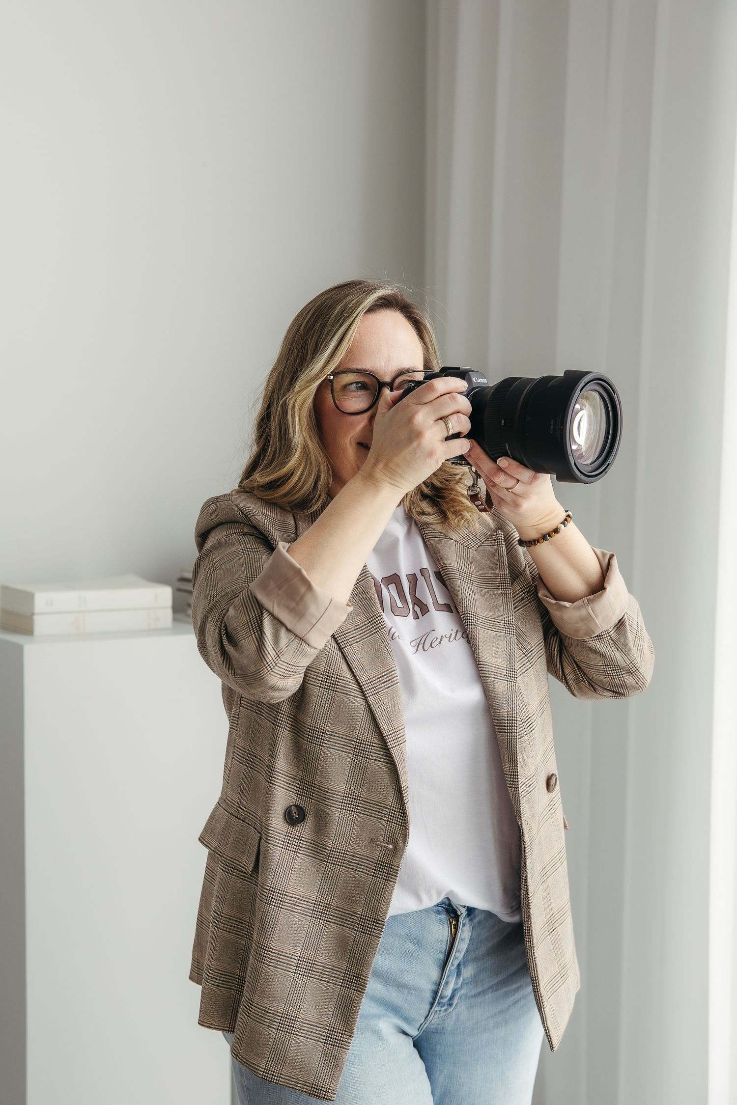 Professional photographer wearing a plaid blazer and jeans takes photos with a DSLR camera near a white wall.