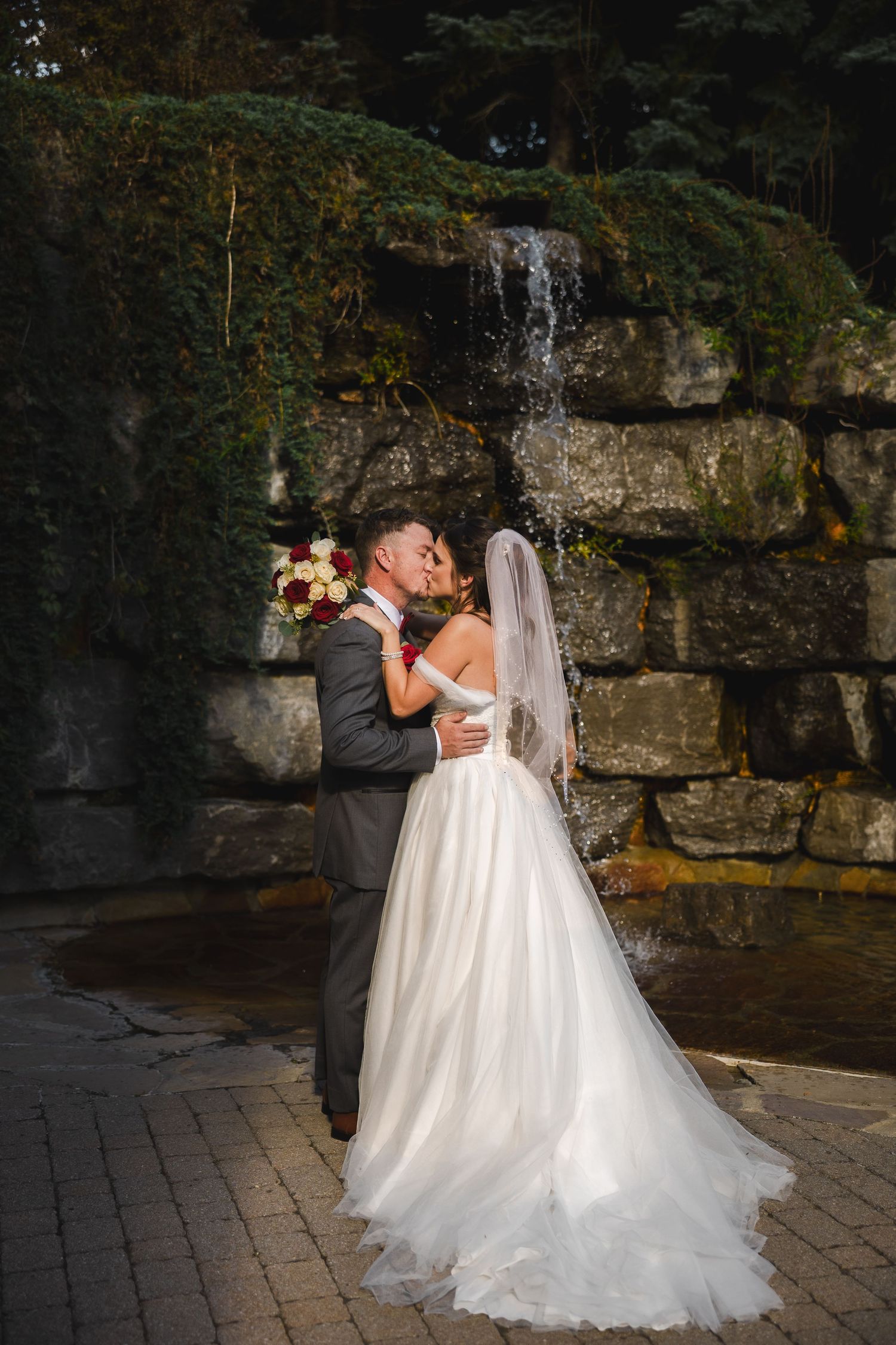 Romantic wedding photos against stone wall and waterfall backdrop at dusk.
