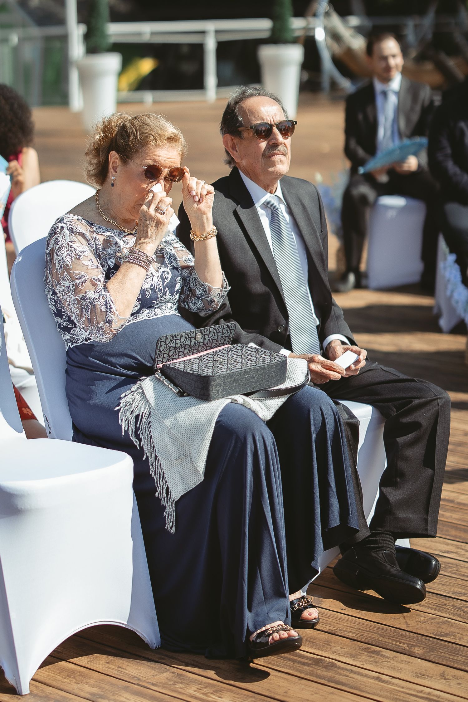 nvités de mariage en tenue élégante assis sur des chaises blanches lors d’une cérémonie extérieure sur une terrasse en bois.