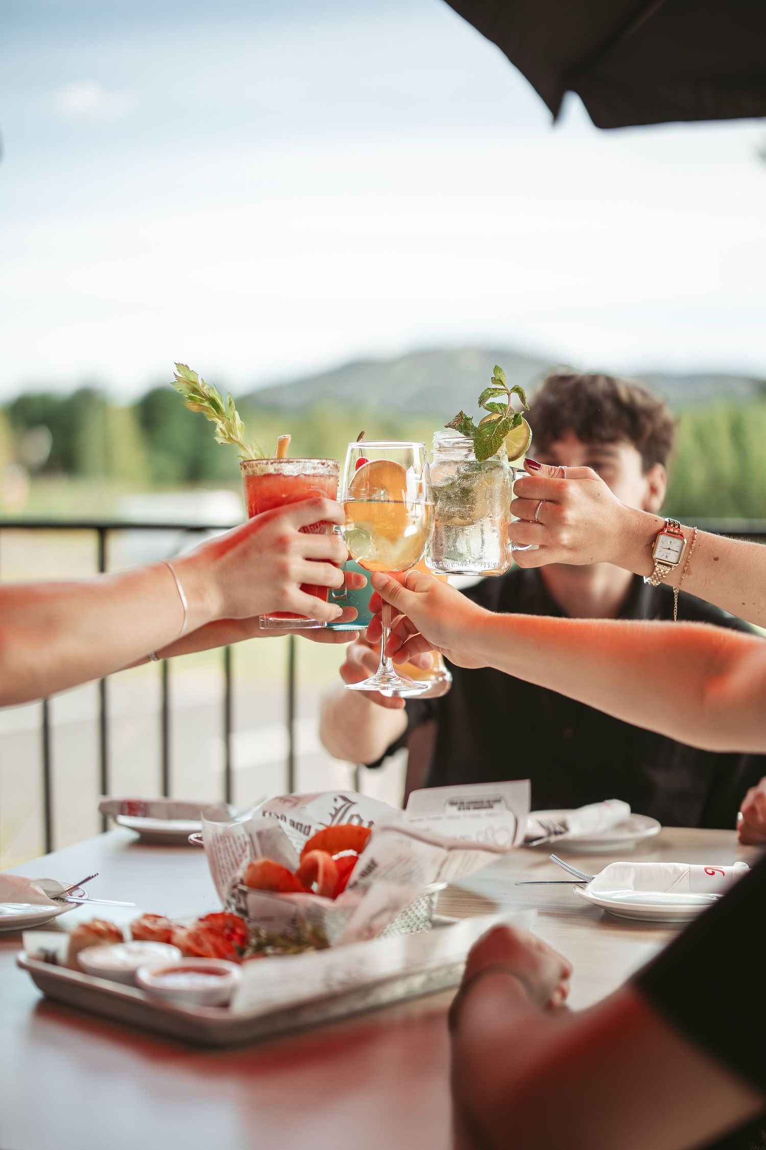 Friends toasting colorful cocktails during an outdoor dining experience with mountain views in the background.