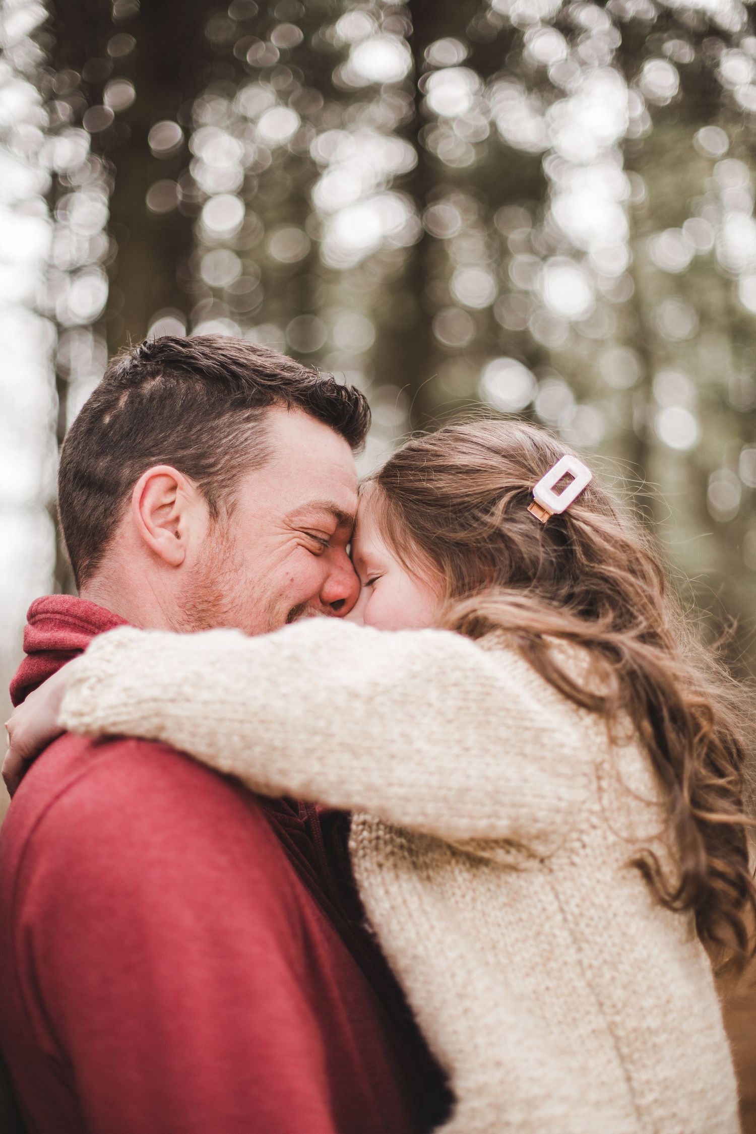 Gros plan : père et fille s’embrassant, pull crème et veste bordeaux, forêt floue en arrière-plan, capturé pour tendresse et complicité