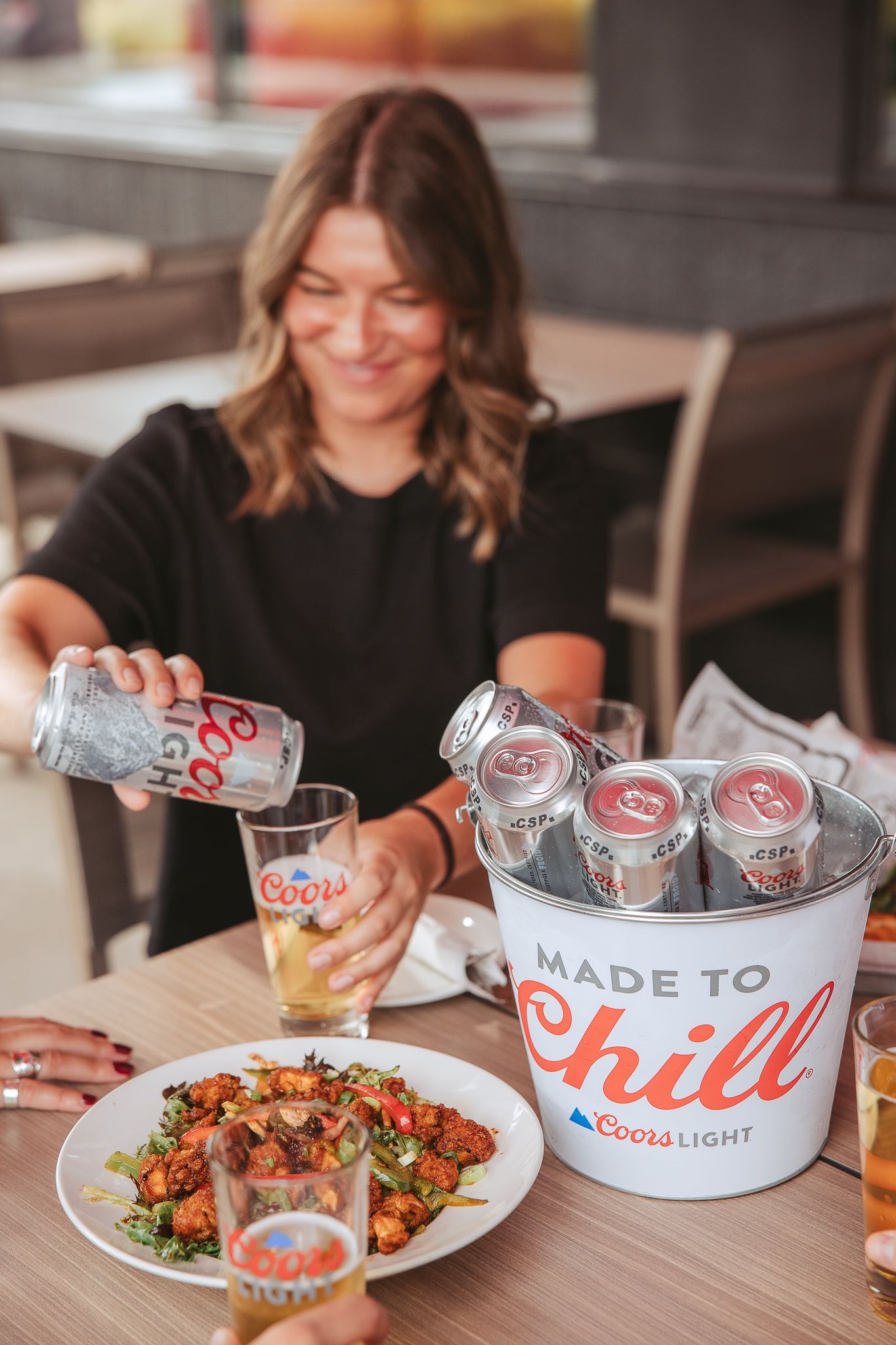 Someone pours a Coors Light beer at a table with food and a branded ice bucket filled with cans.