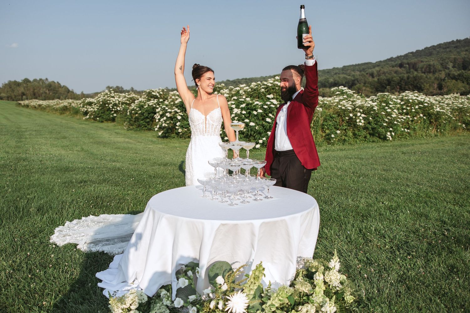 Mariée en robe blanche levant les bras de joie lors d’un mariage en plein air, avec collines verdoyantes en arrière-plan.