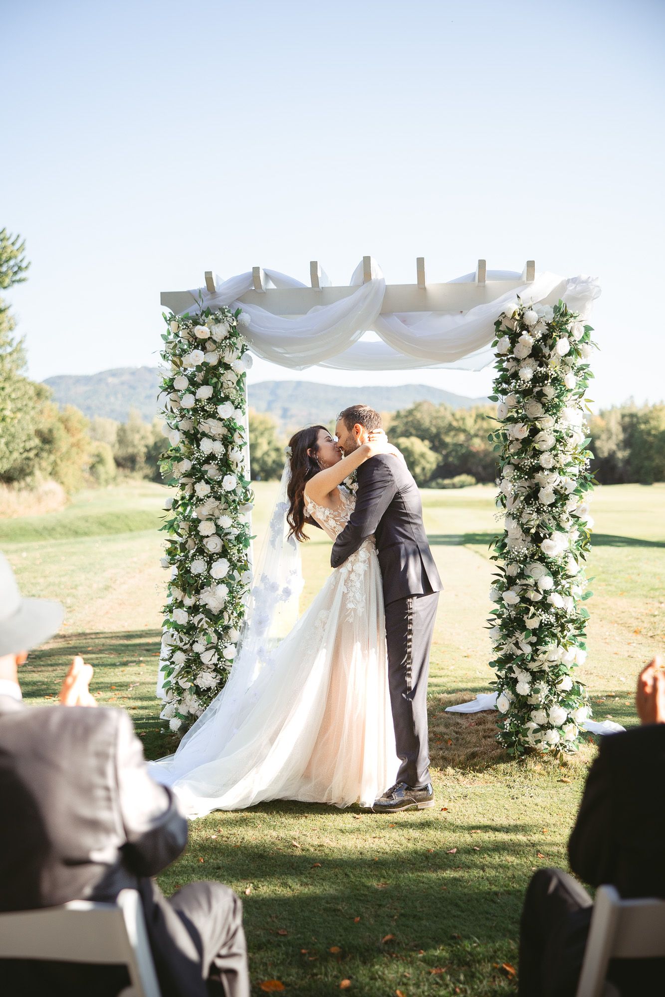 Cérémonie de mariage en plein air sous une arche blanche drapée et fleurie, avec montagnes en arrière-plan à Bromont