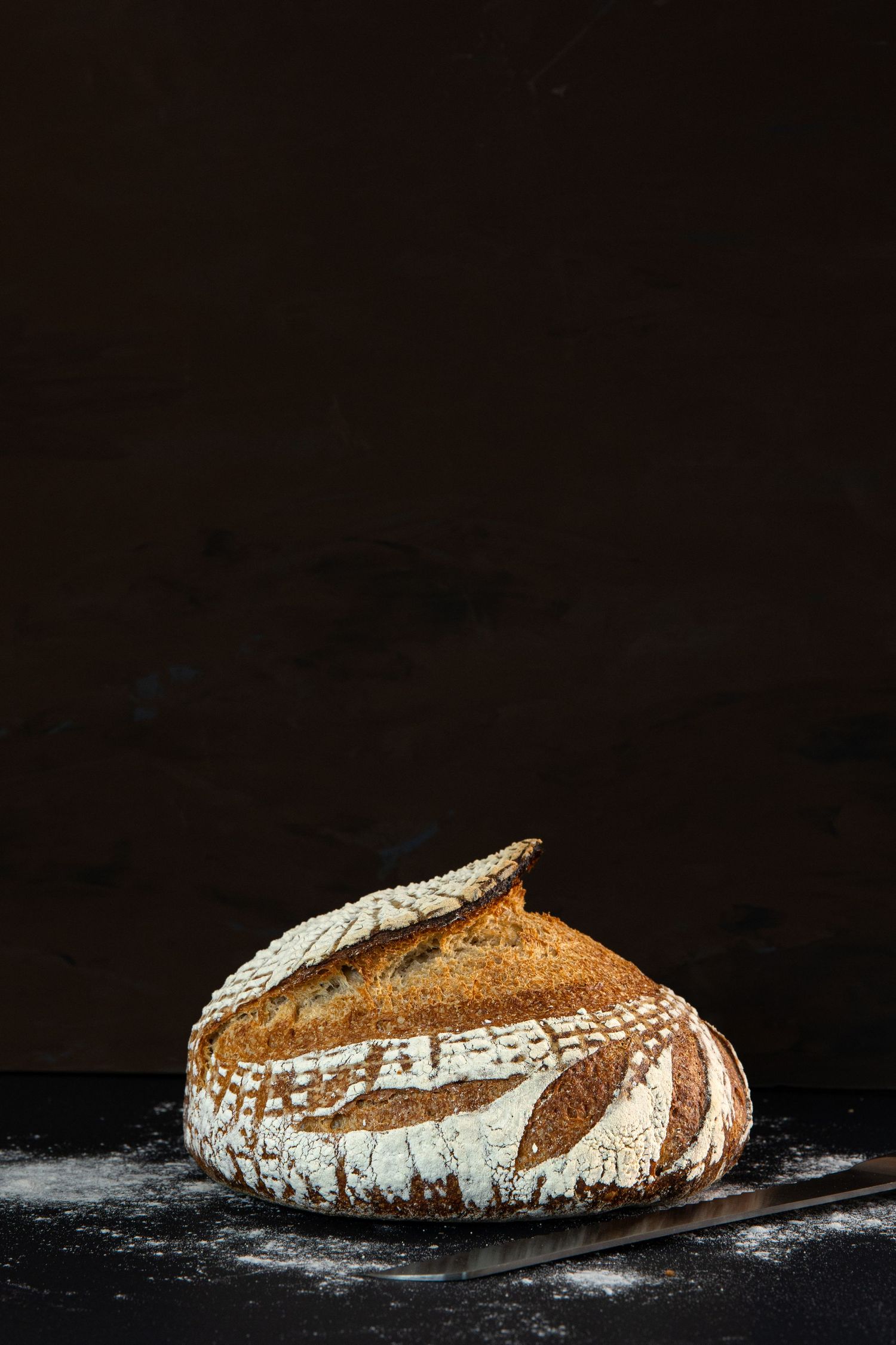 Artisanal sourdough bread with scored pattern and flour dusting on black surface.