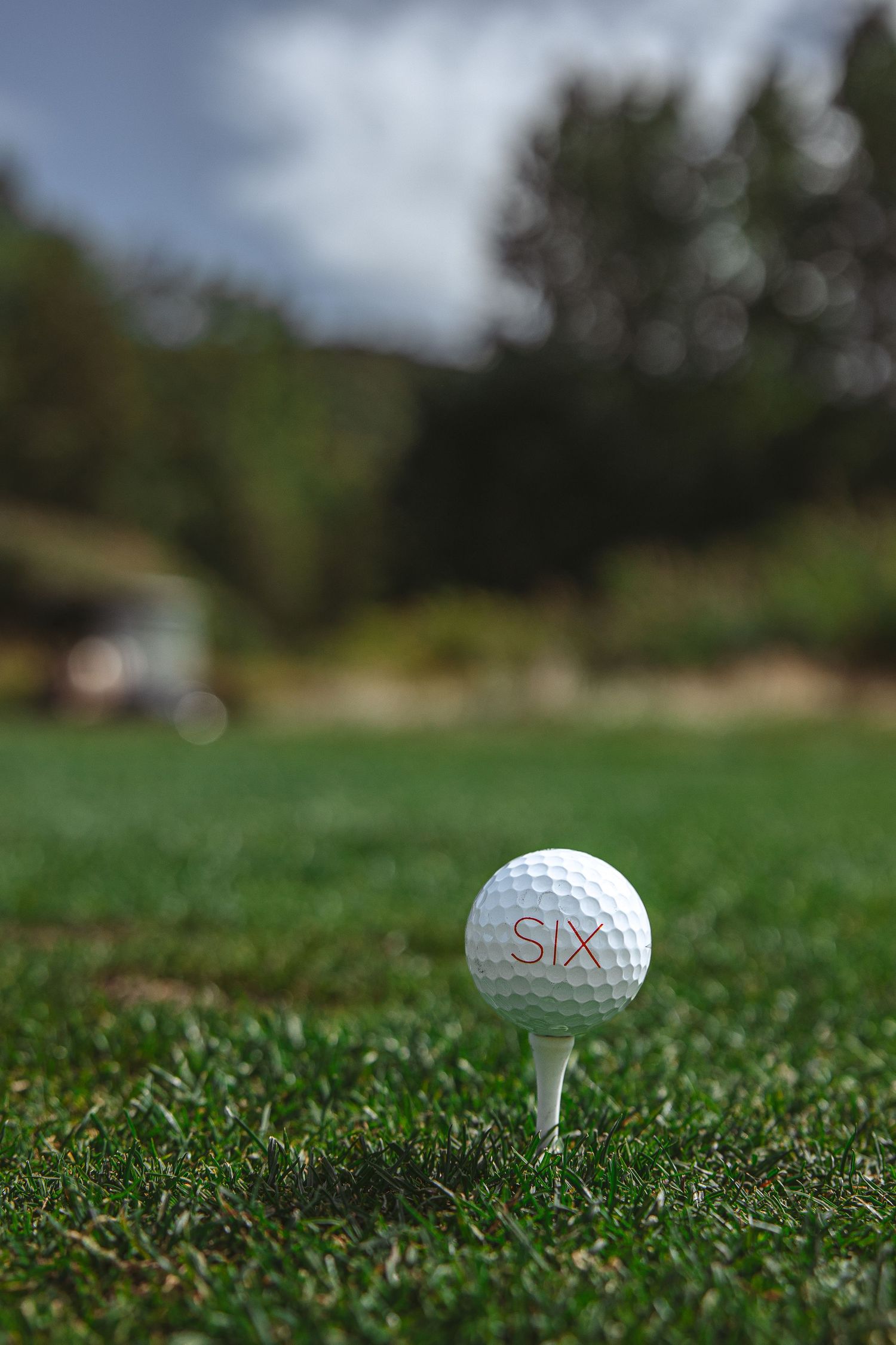 White golf ball on a tee sits in lush green grass on a golf course under cloudy skies.
