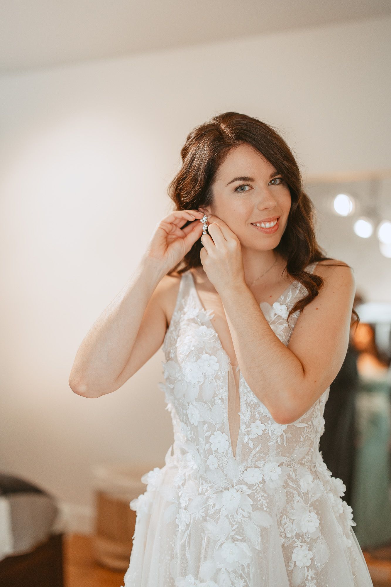 Mariée en robe blanche à appliqués floraux délicats mettant ses boucles d’oreilles pendant les préparatifs