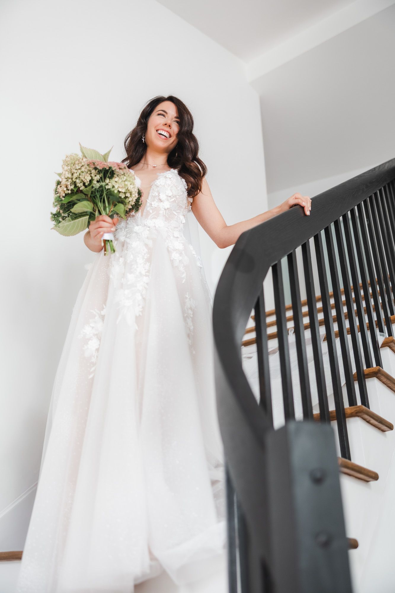 Mariée en robe blanche fluide en dentelle descendant un escalier à rampe noire en riant, mariage en Estrie
