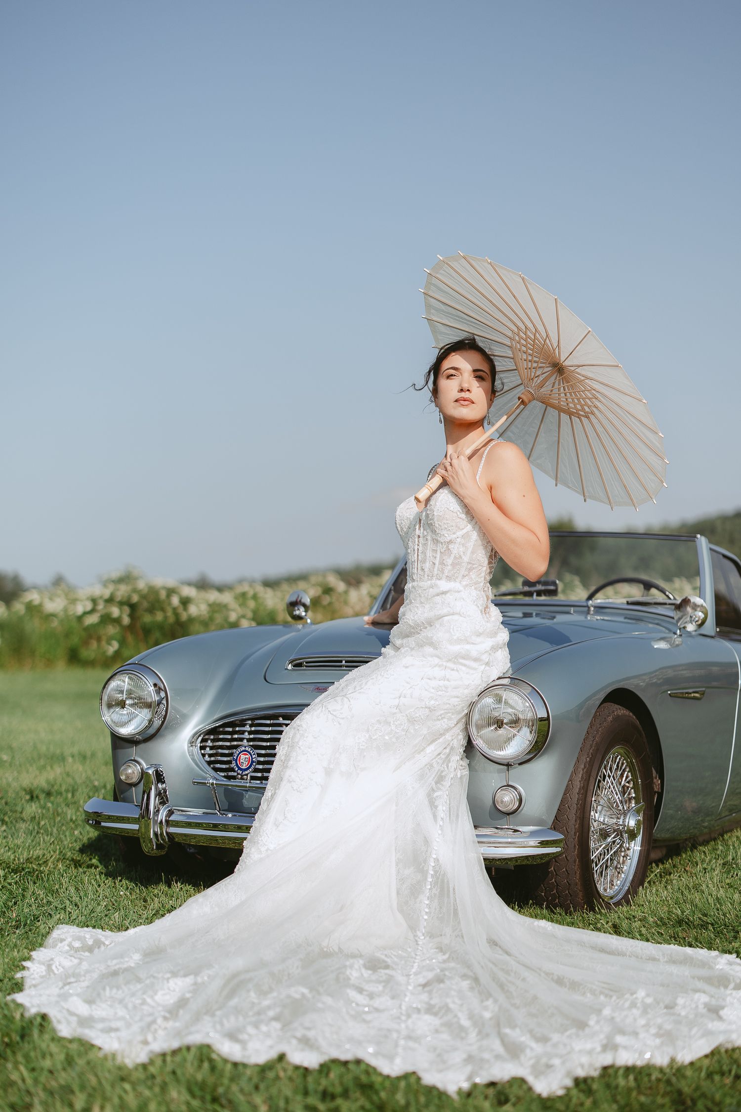 Série photo de mariage mettant en scène une voiture décapotable Austin vintage, une robe blanche fluide et une ombrelle en extérieur.